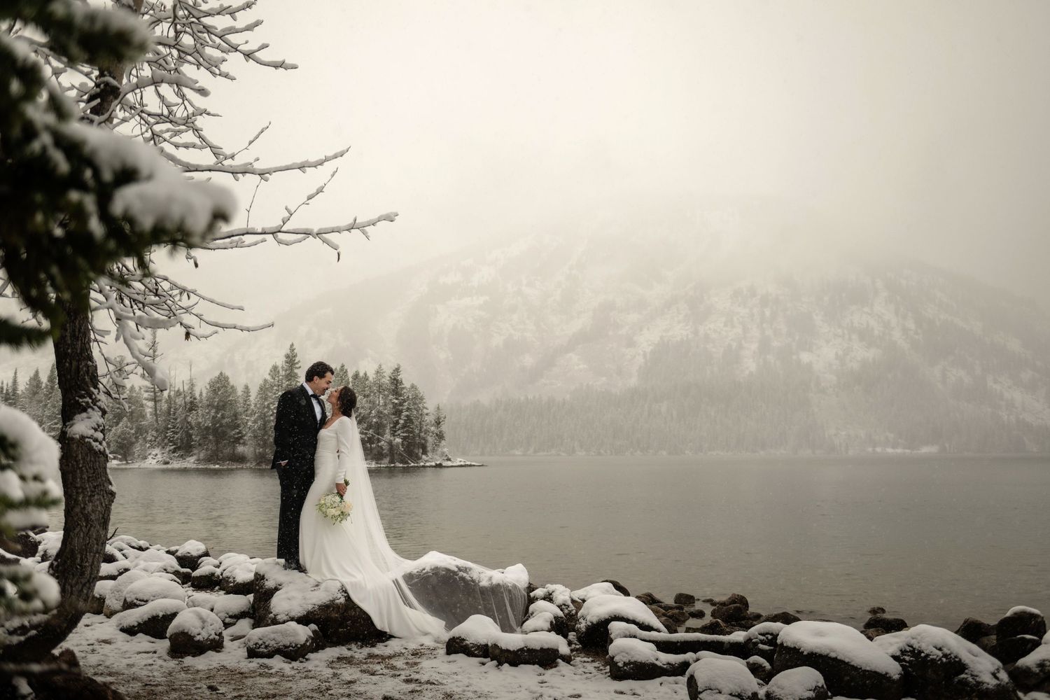Bride and groom share a romantic winter kiss by a snow-covered mountain lake surrounded by frosted evergreen trees.