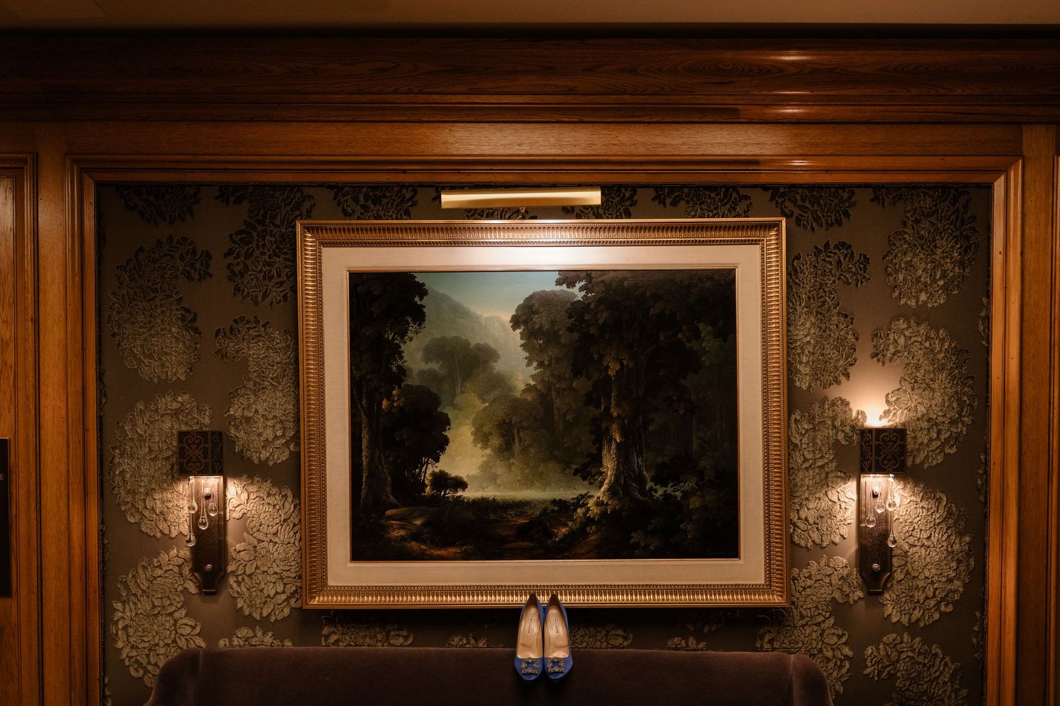 A framed landscape painting hangs on the wall of a cozy, dimly lit room with sconces.
