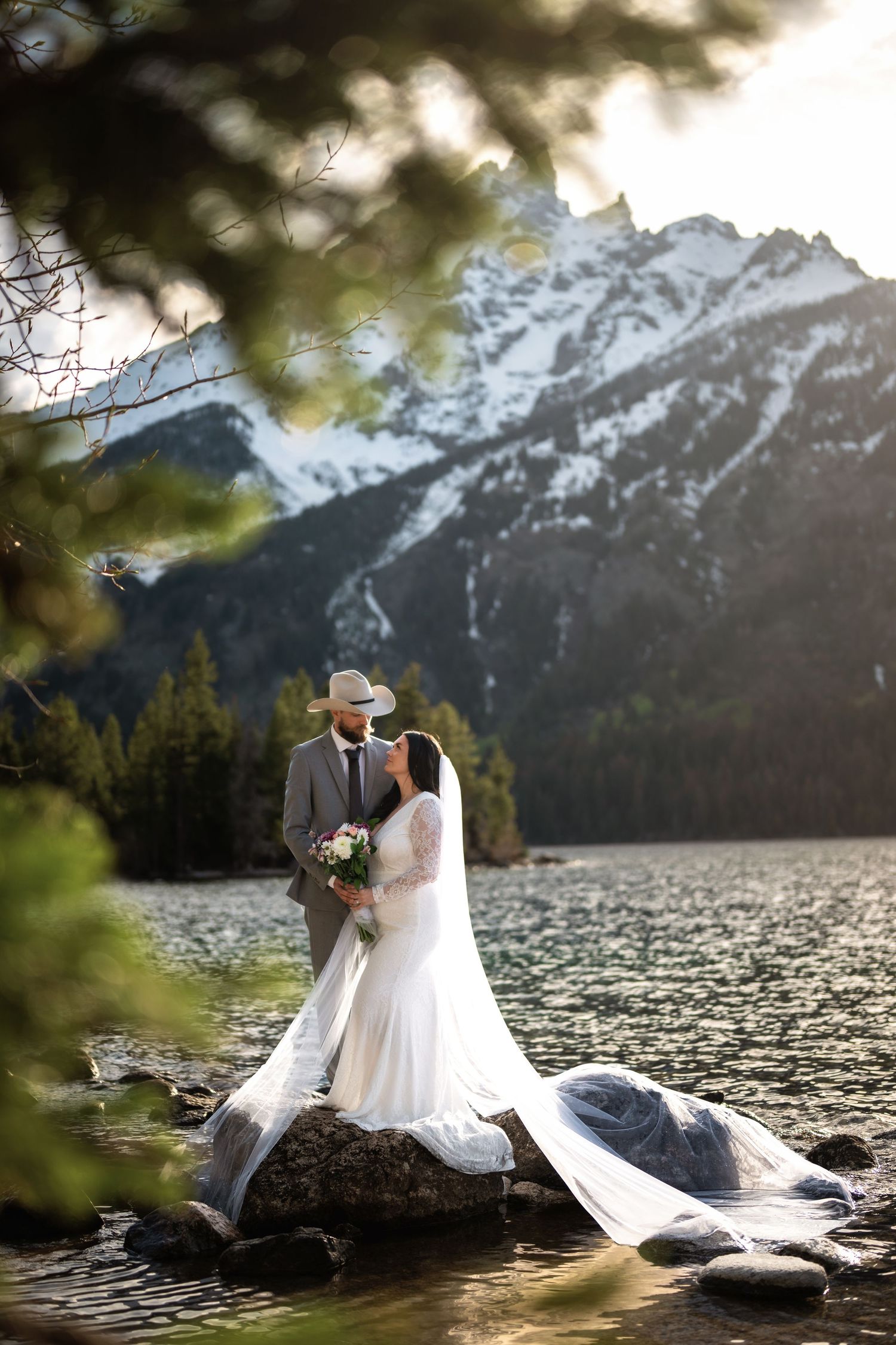Bride and groom kissing on rocks by a mountain lake with snowy peaks and pine trees in the background.