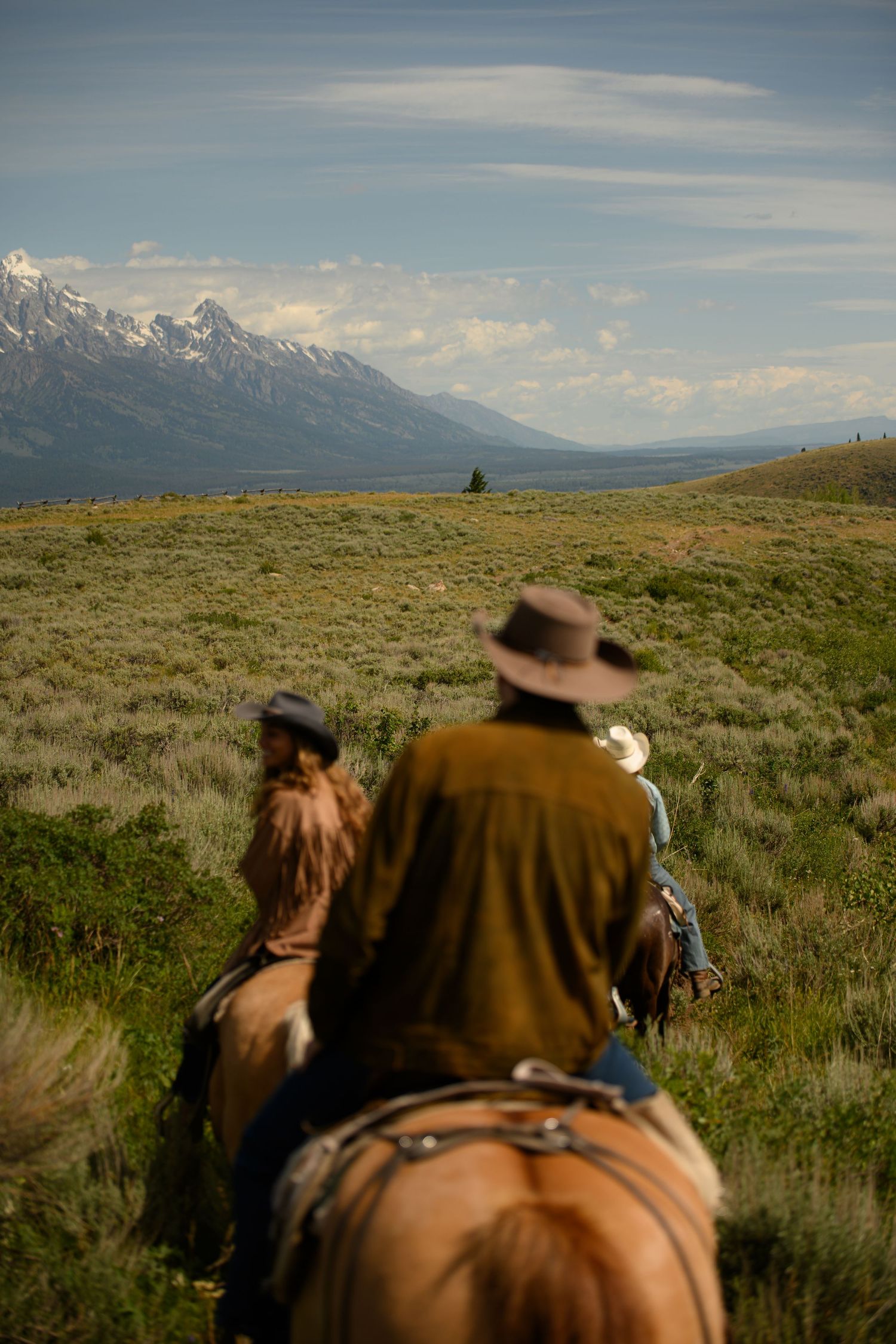 Two cowboys riding horses through green meadows with snow-capped Grand Teton mountains in the background.
