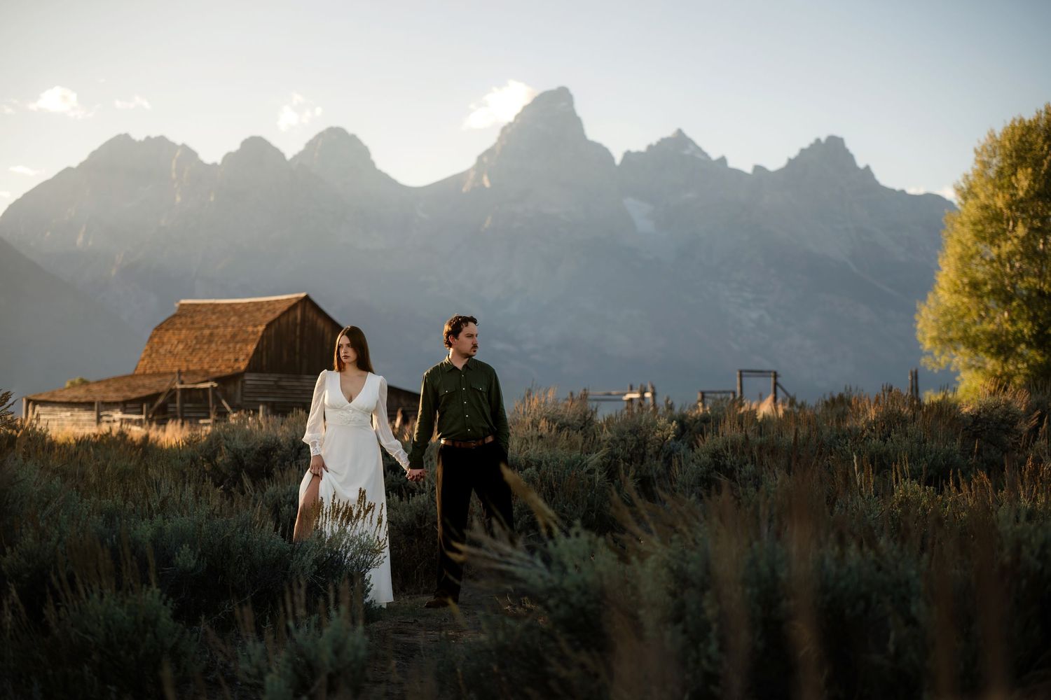 Couple holds hands in front of rustic barn with Grand Teton mountains at golden hour in Jackson Hole, Wyoming.