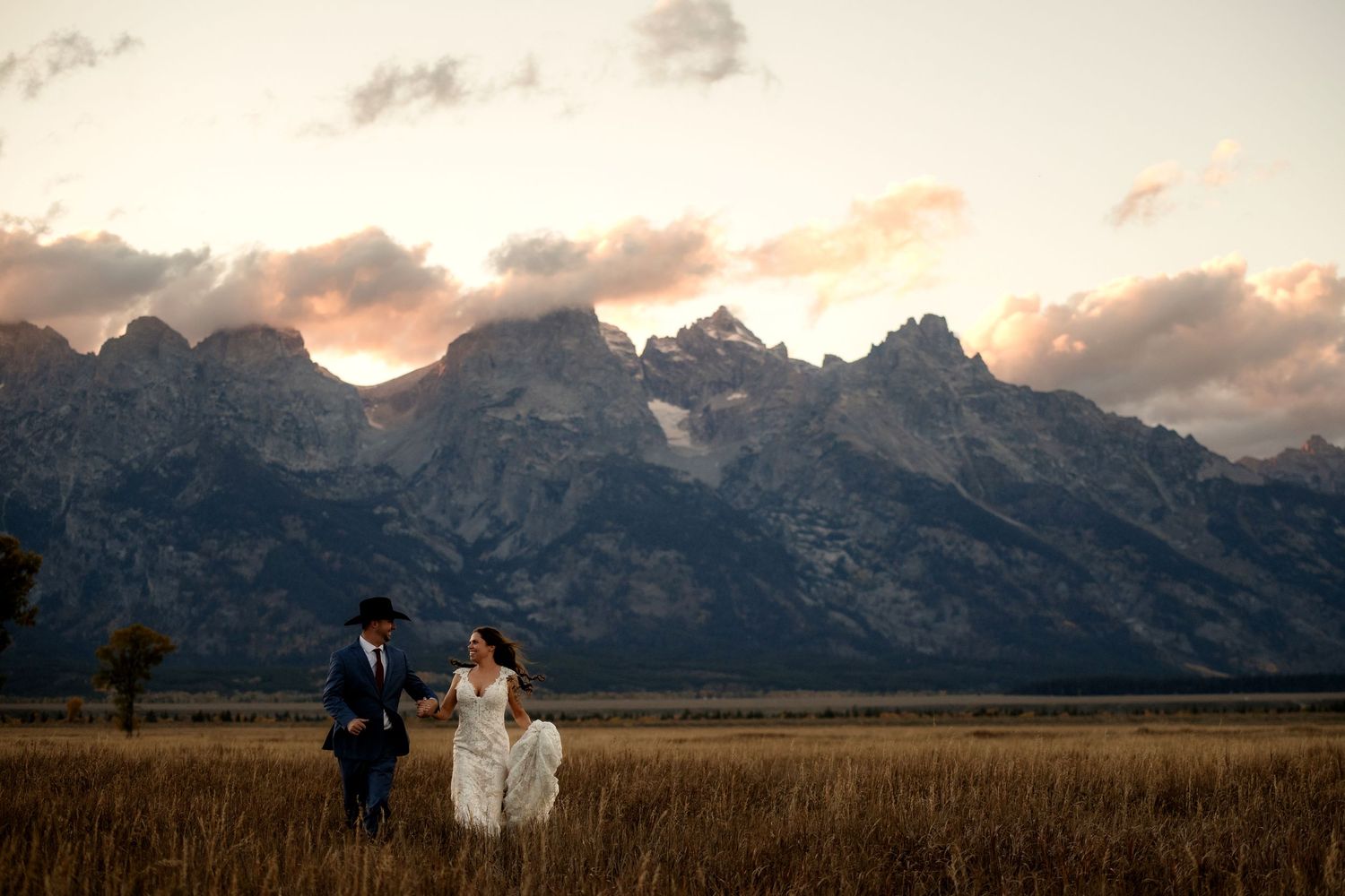 A bride and groom walk through a field with mountains in the background.