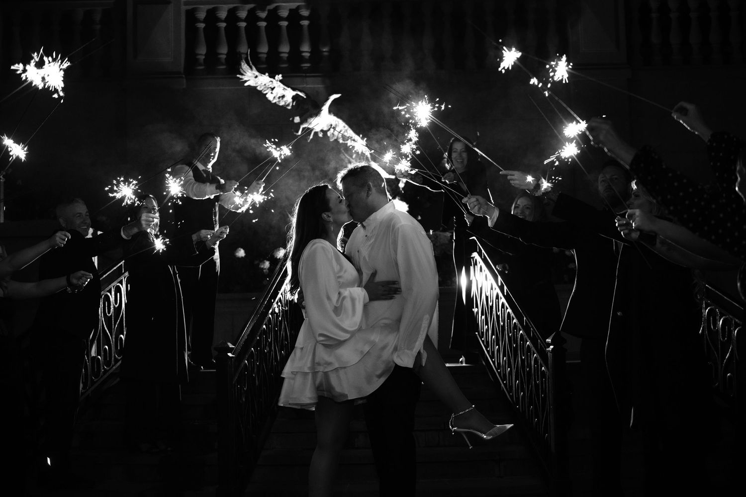 Black and white photo of a couple kissing surrounded by sparklers at night during a wedding send-off.
