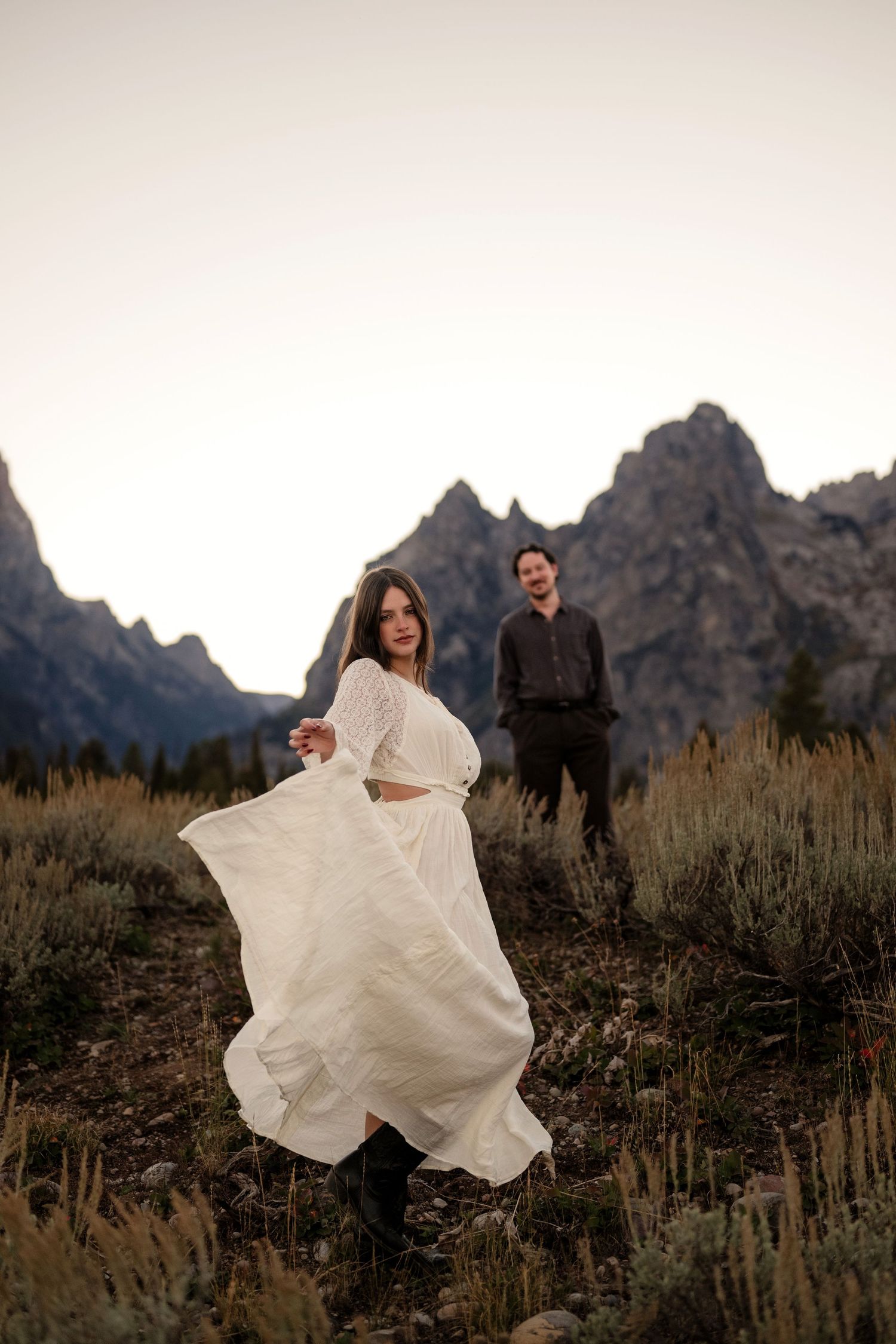 Woman in white flowing dress twirls before rugged mountain peaks at dusk with man standing in background.