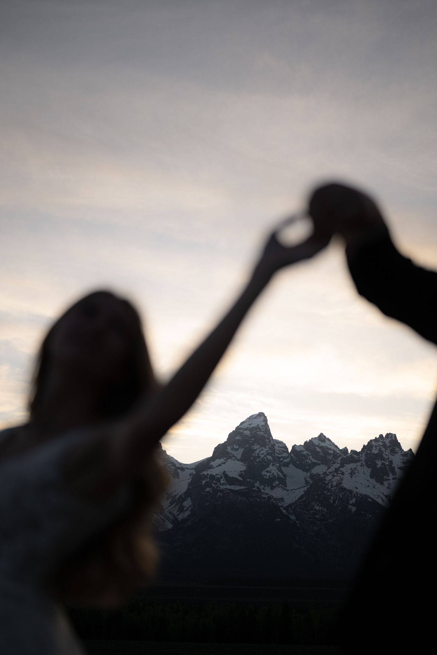 Silhouette of couple holding hands raised against snow-capped mountain peaks at dusk.