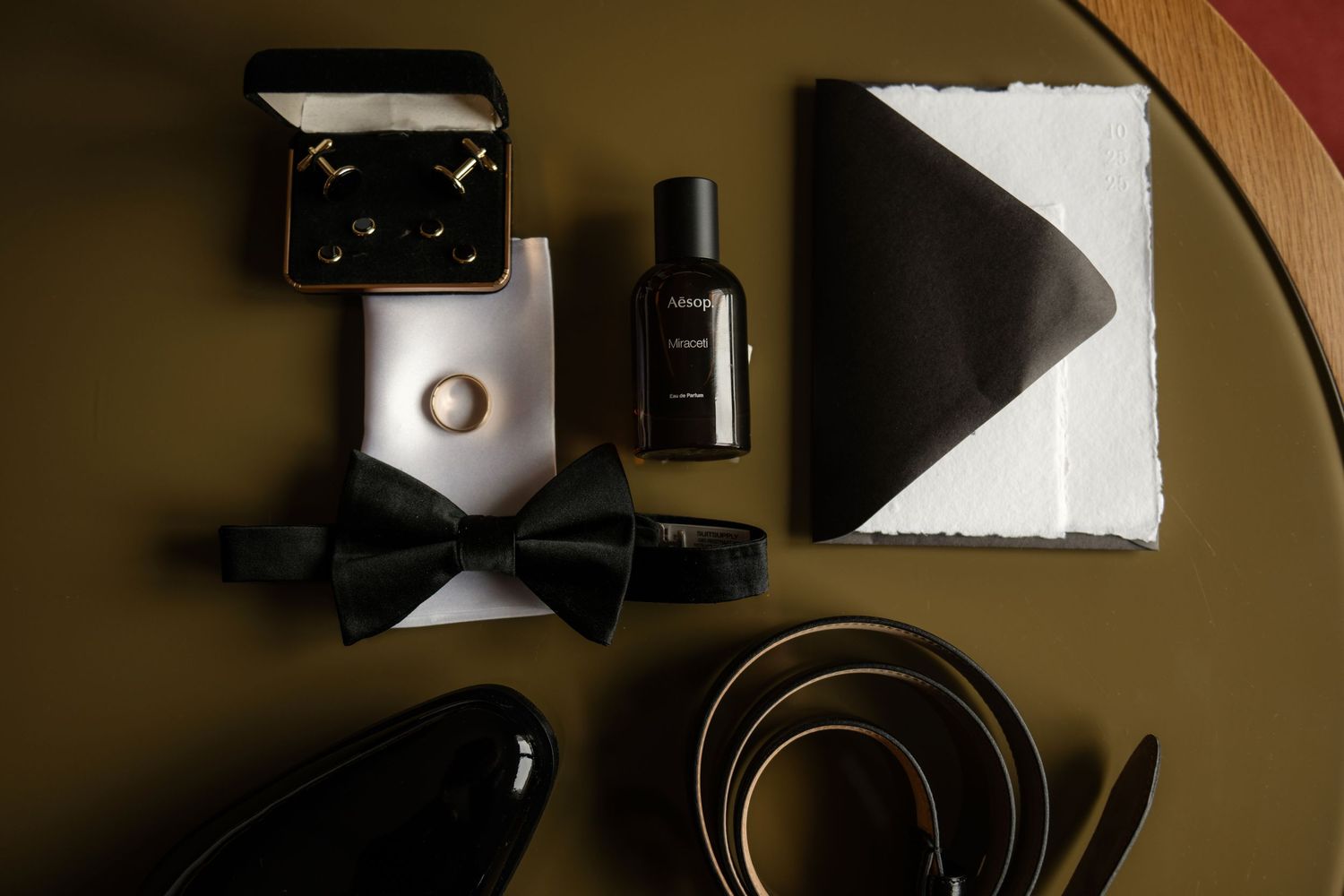 Groom wedding flat lay with bow tie, cologne, cufflinks, belt, and shoes on olive green surface.