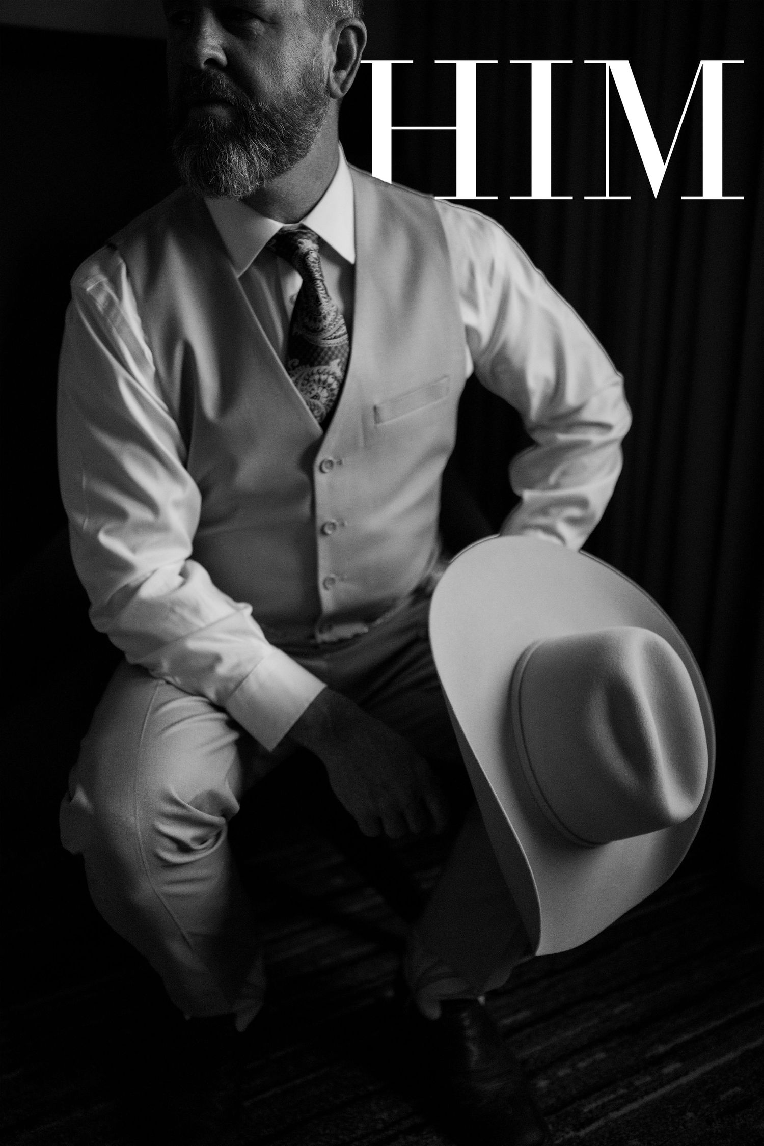 Black and white photo of bearded man in vest and tie holding cowboy hat, styled for HIM magazine editorial.