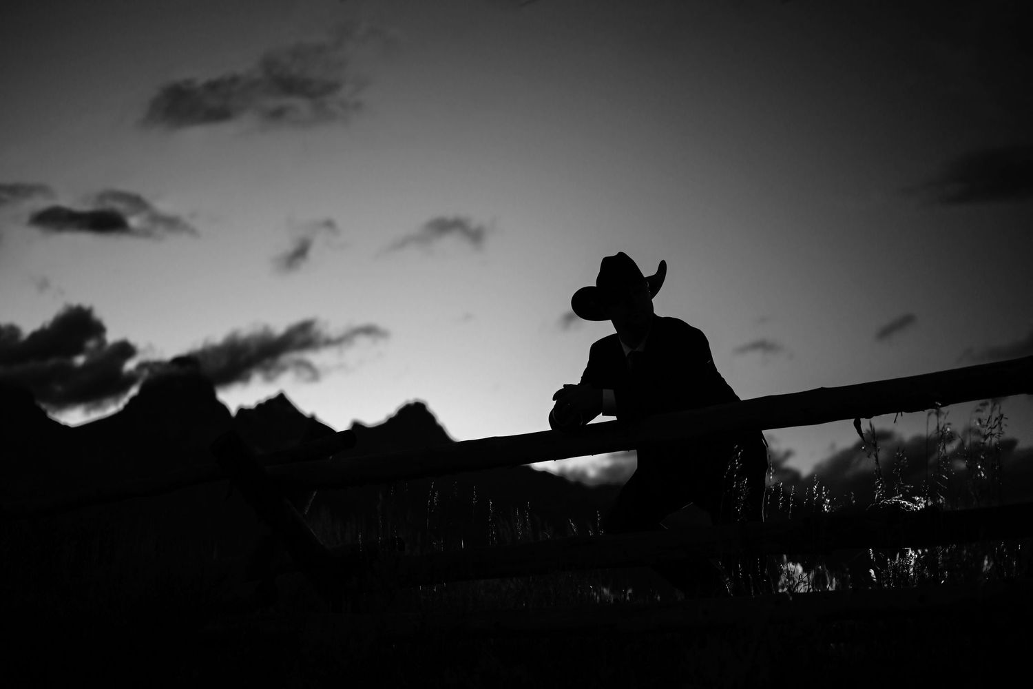 Silhouette of cowboy leaning on fence rail against dramatic sunset sky, black and white.