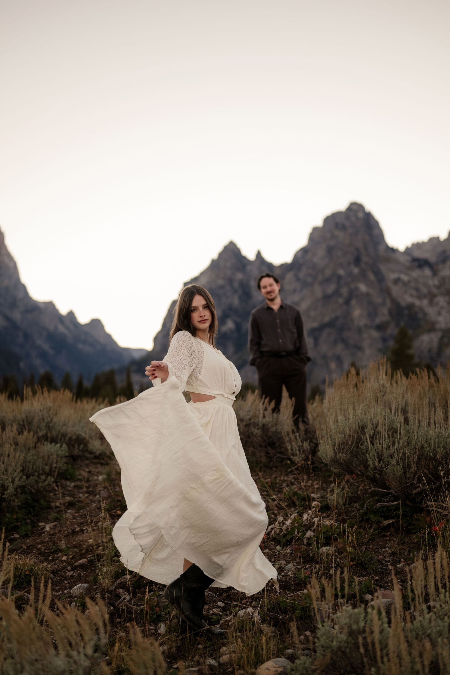 Woman in white flowing dress twirls before rugged mountain backdrop at dusk, man in dark attire stands behind her.