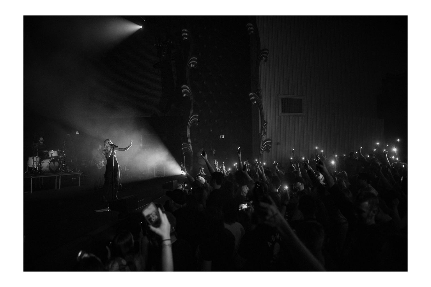 Black and white concert photo showing performer on stage with crowd holding up phone lights in dark venue.
