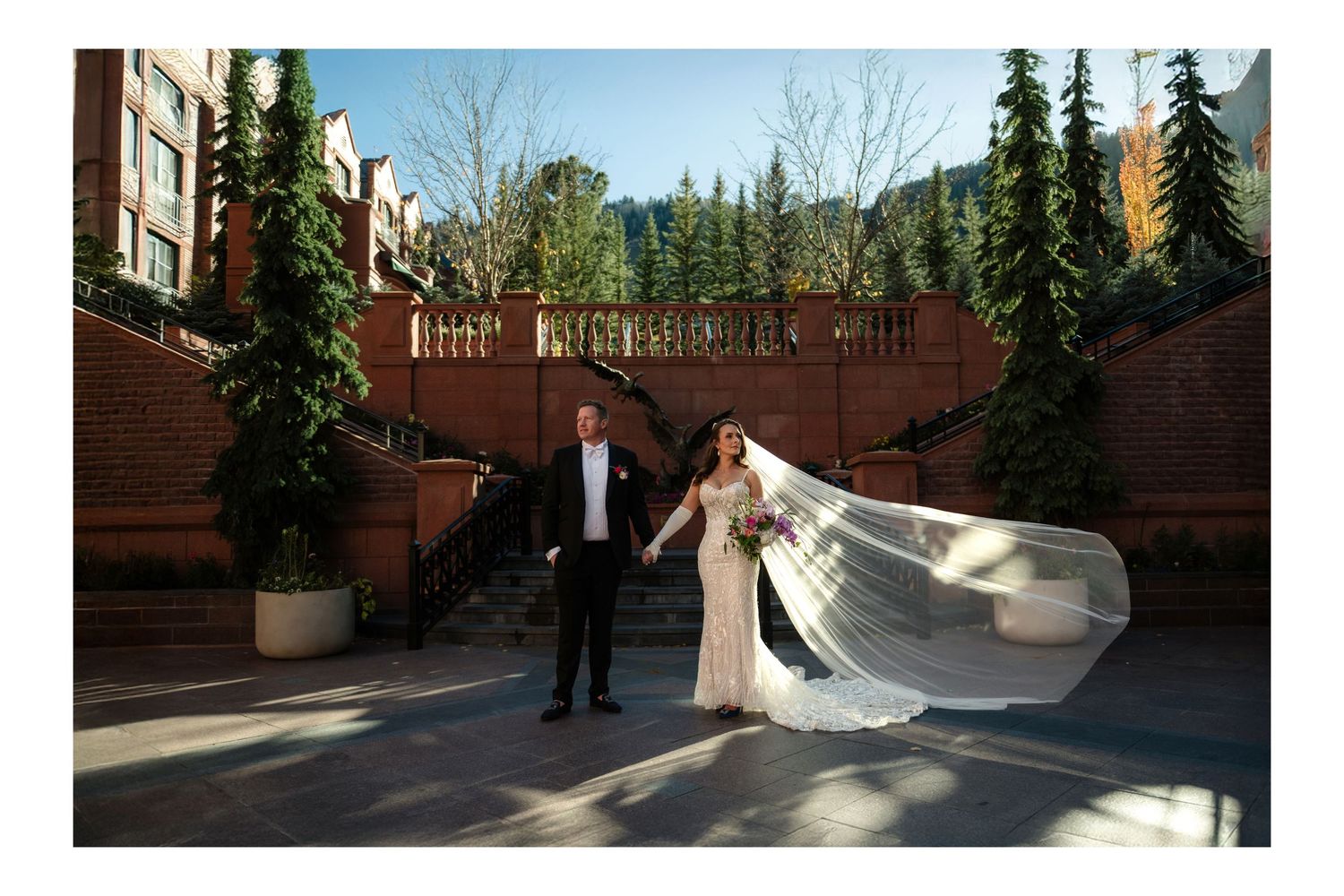 Bride and groom holding hands outdoors at a brick wedding venue with evergreen trees.