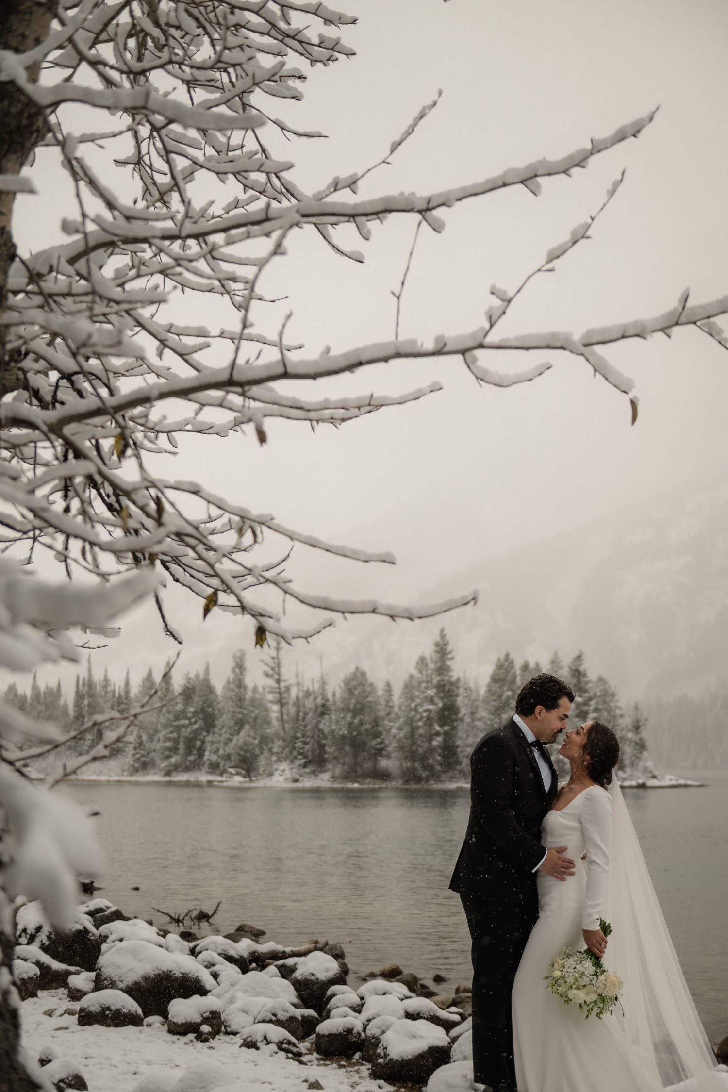 Bride and groom share a kiss in a snowy winter landscape beside a lake, surrounded by snow-covered trees and rocks.
