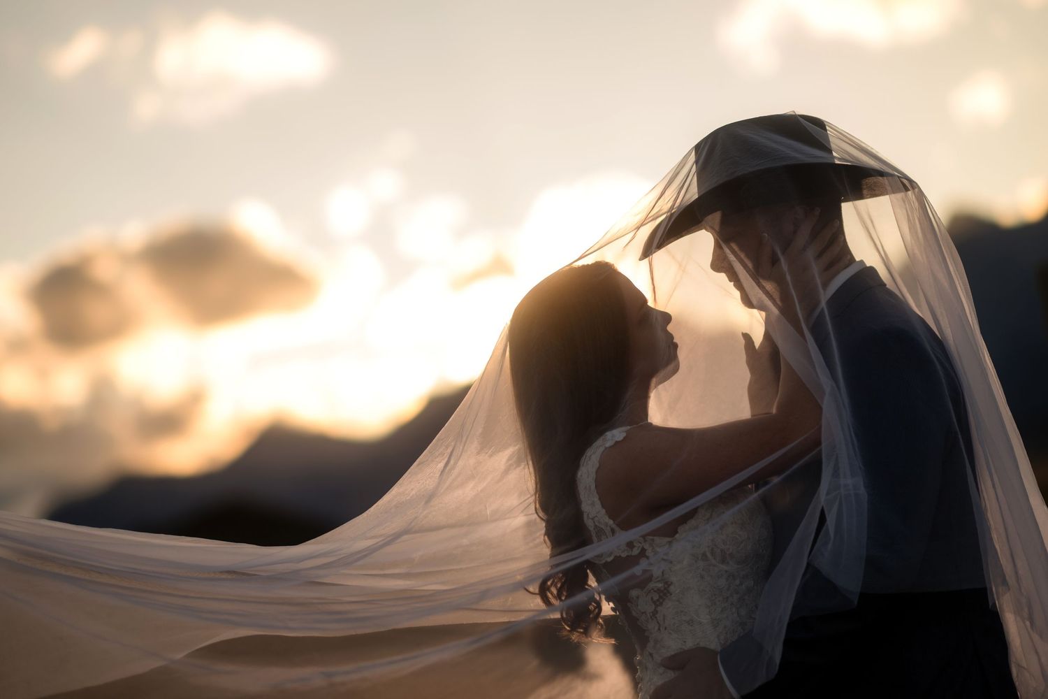 Bride and groom share a romantic sunset moment under a flowing veil with mountains in the background.
