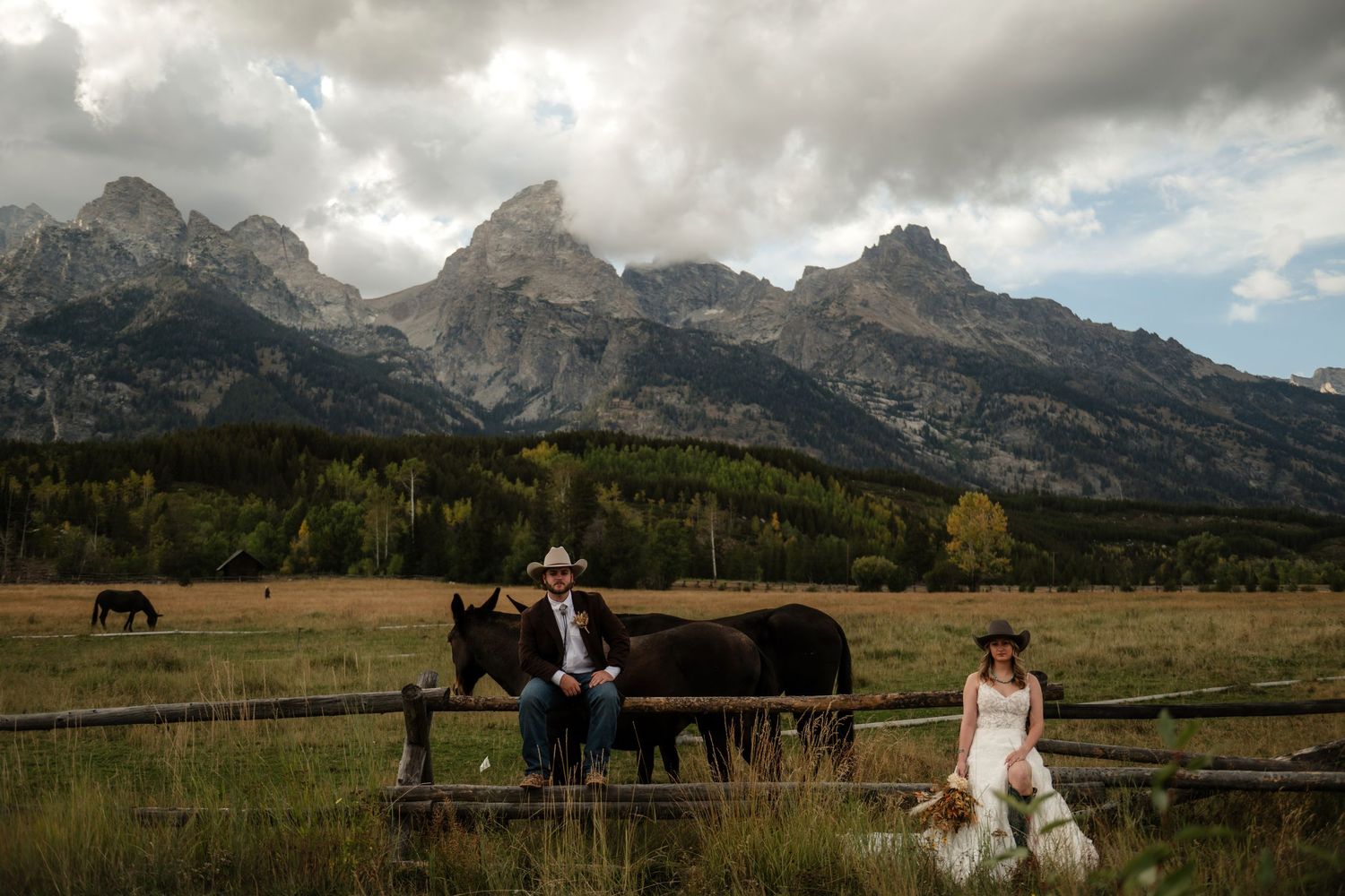 Couple in western attire sits on a fence with horses, framed by the dramatic Grand Teton mountains under cloudy skies.