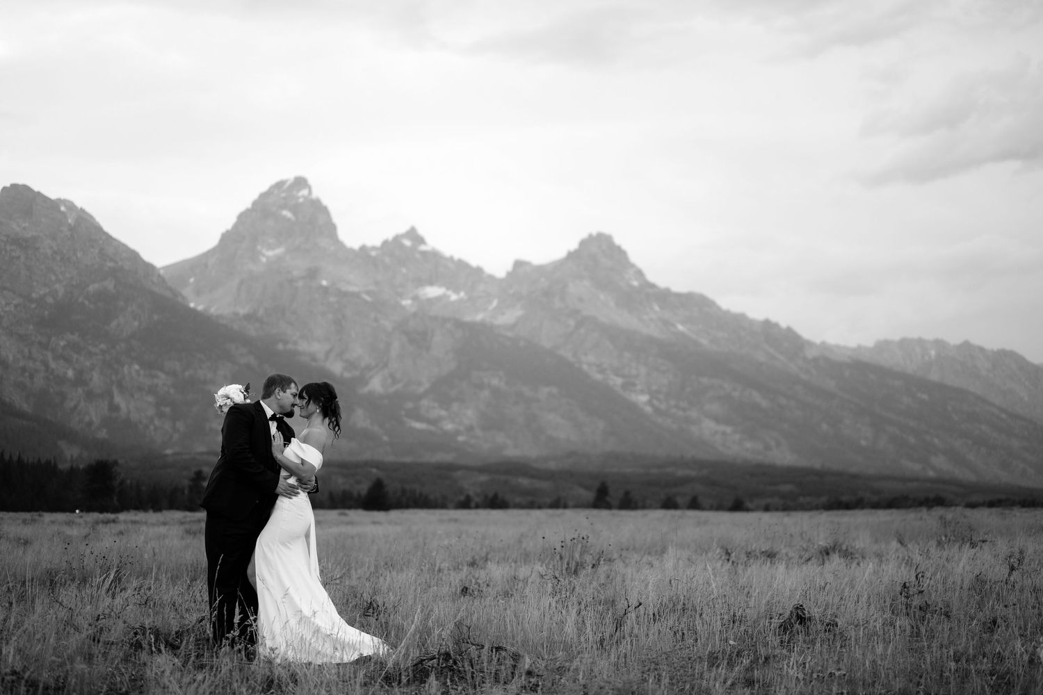 Black and white photo of a bride and groom embracing in a meadow with the Grand Teton mountains in the background.