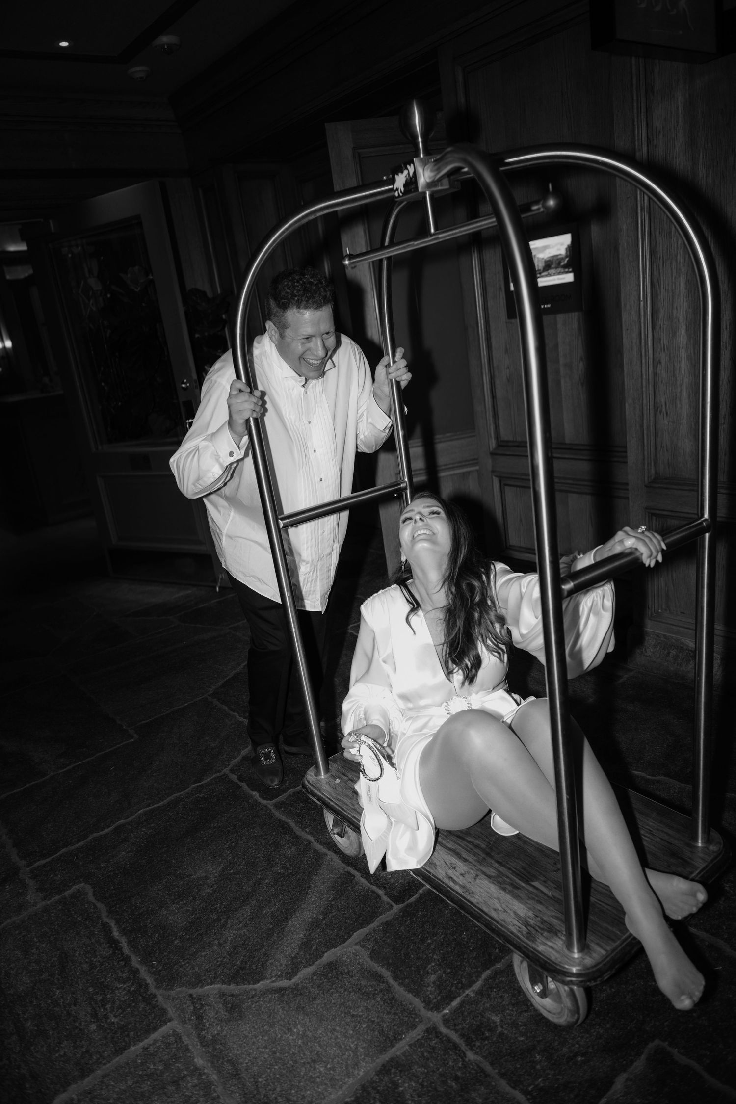Black and white photo of a laughing couple playing on a hotel luggage cart in a rustic lobby.