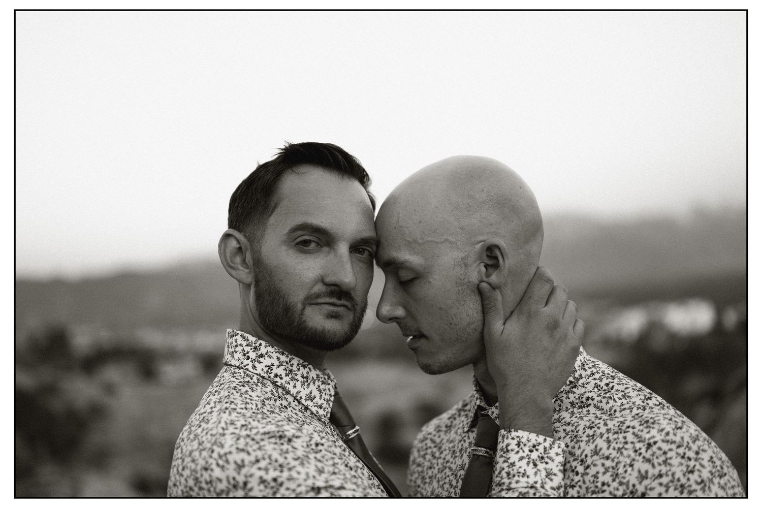 Black and white portrait of two men sharing an intimate moment outdoors, one tenderly cradling the others face.