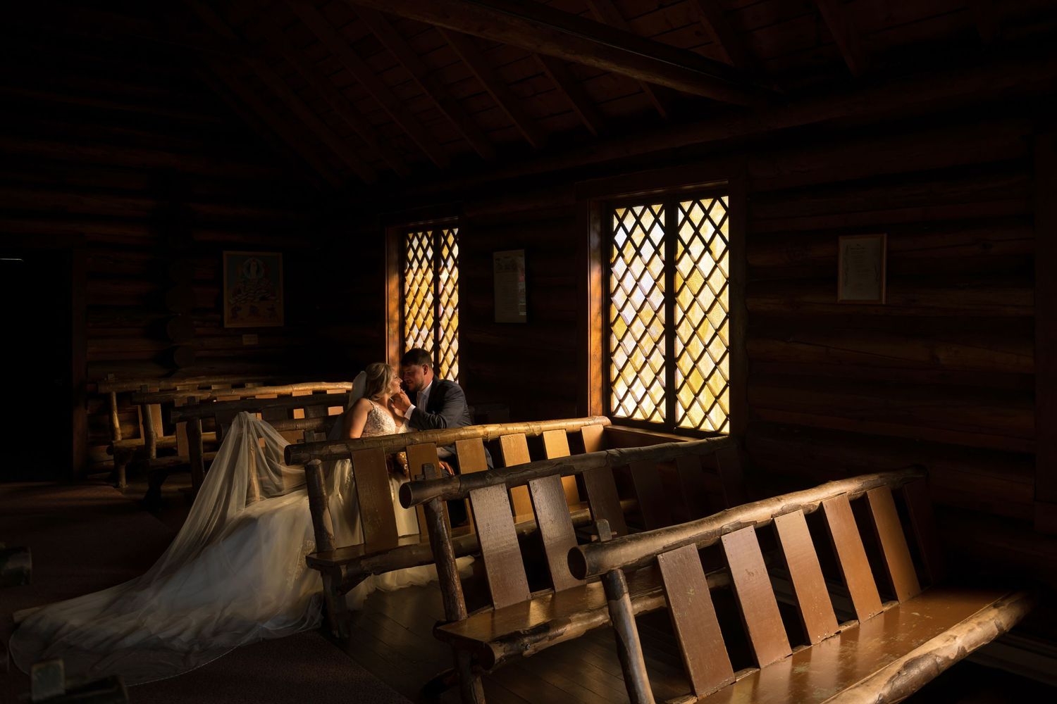 Bride and groom sit together on wooden church pews bathed in warm golden light from latticed windows in a rustic log chapel.