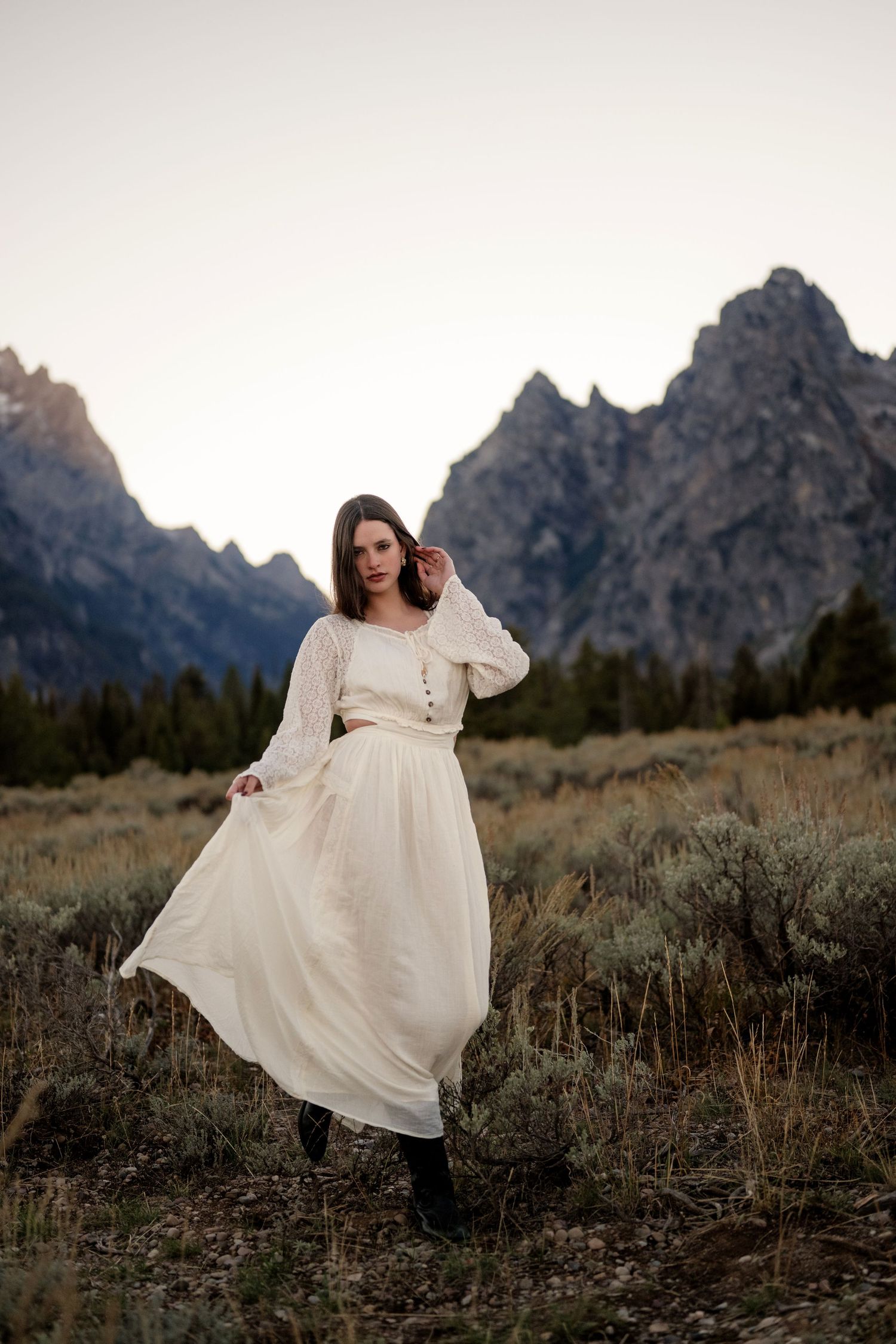 Woman in flowing cream dress poses in sagebrush field with Teton mountains at dusk.