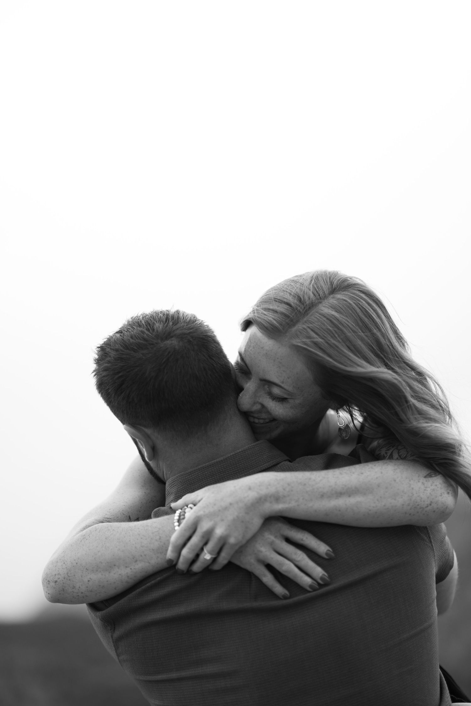 Black and white photo of a smiling woman hugging a man outdoors, wearing an engagement ring.
