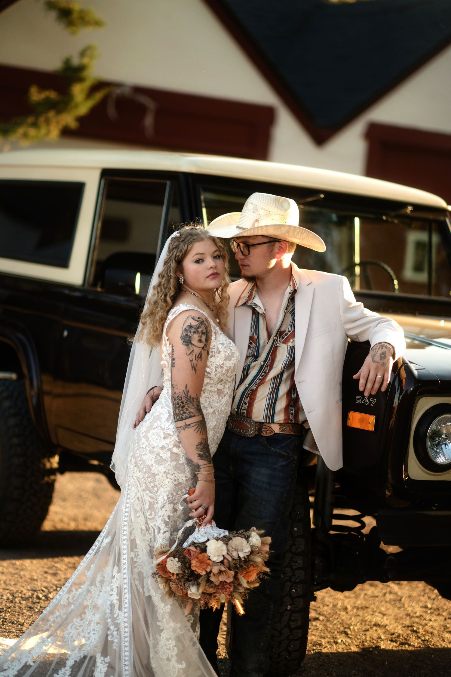A black and white image shows a bride and groom embracing in front of a vintage car.