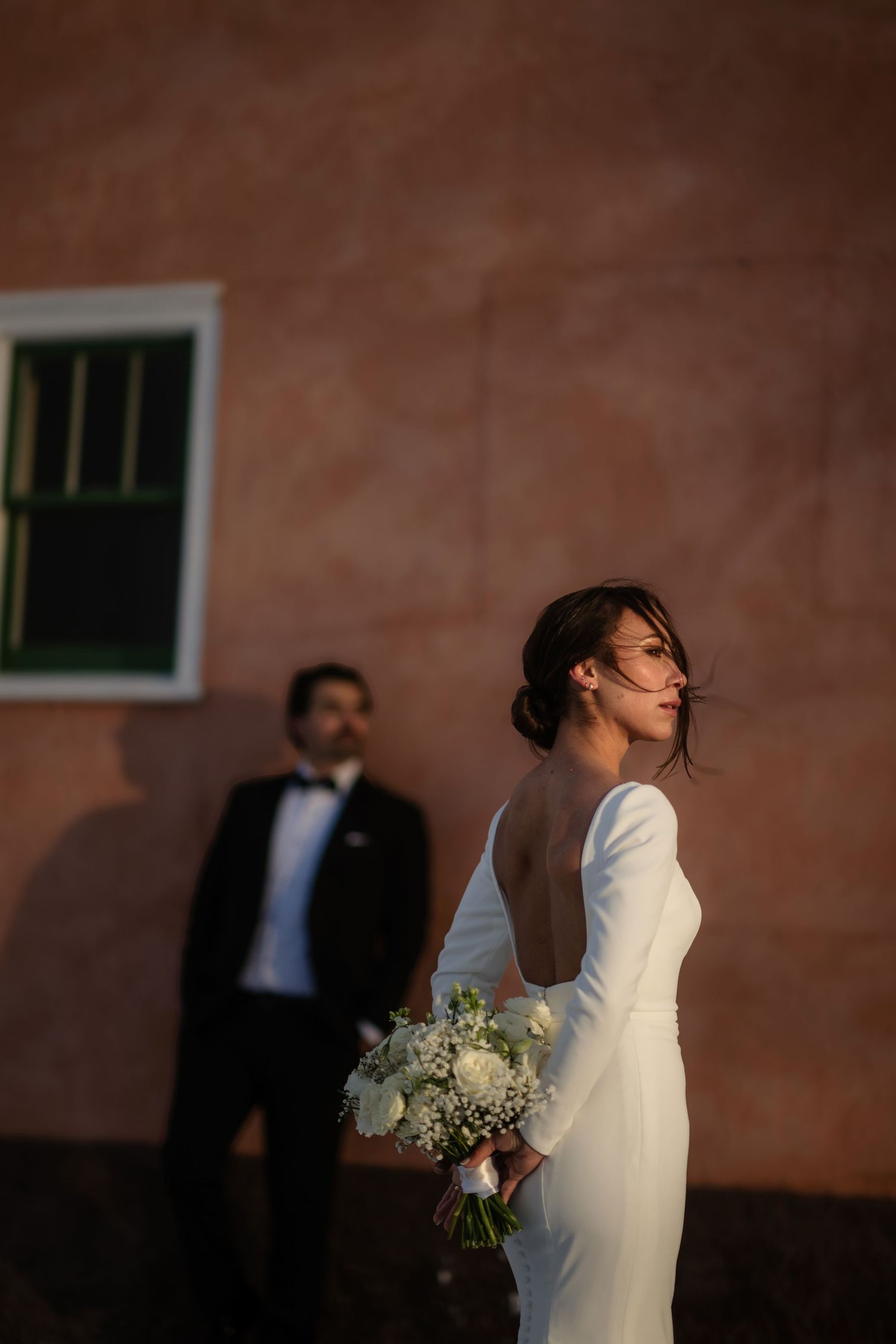 Bride in backless white gown holds bouquet while groom in tuxedo stands against a warm terracotta wall.