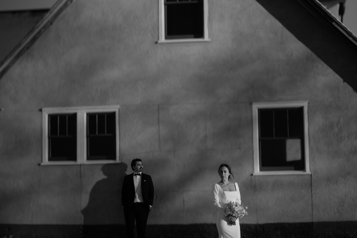 Black and white wedding photo of bride and groom standing apart against a sunlit building wall.