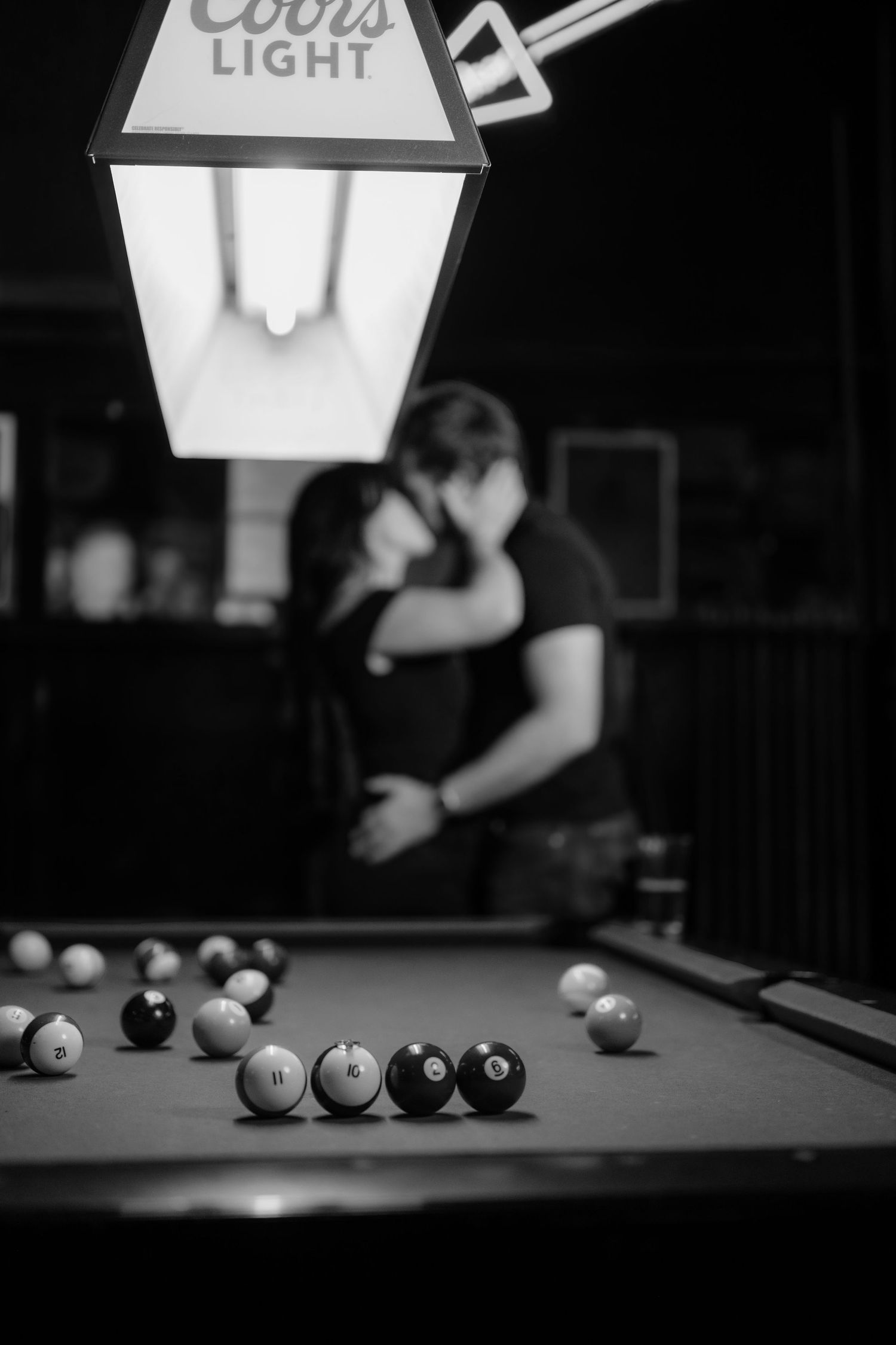 Black and white photo of couple kissing near a pool table under a Coors Light lamp in a bar.