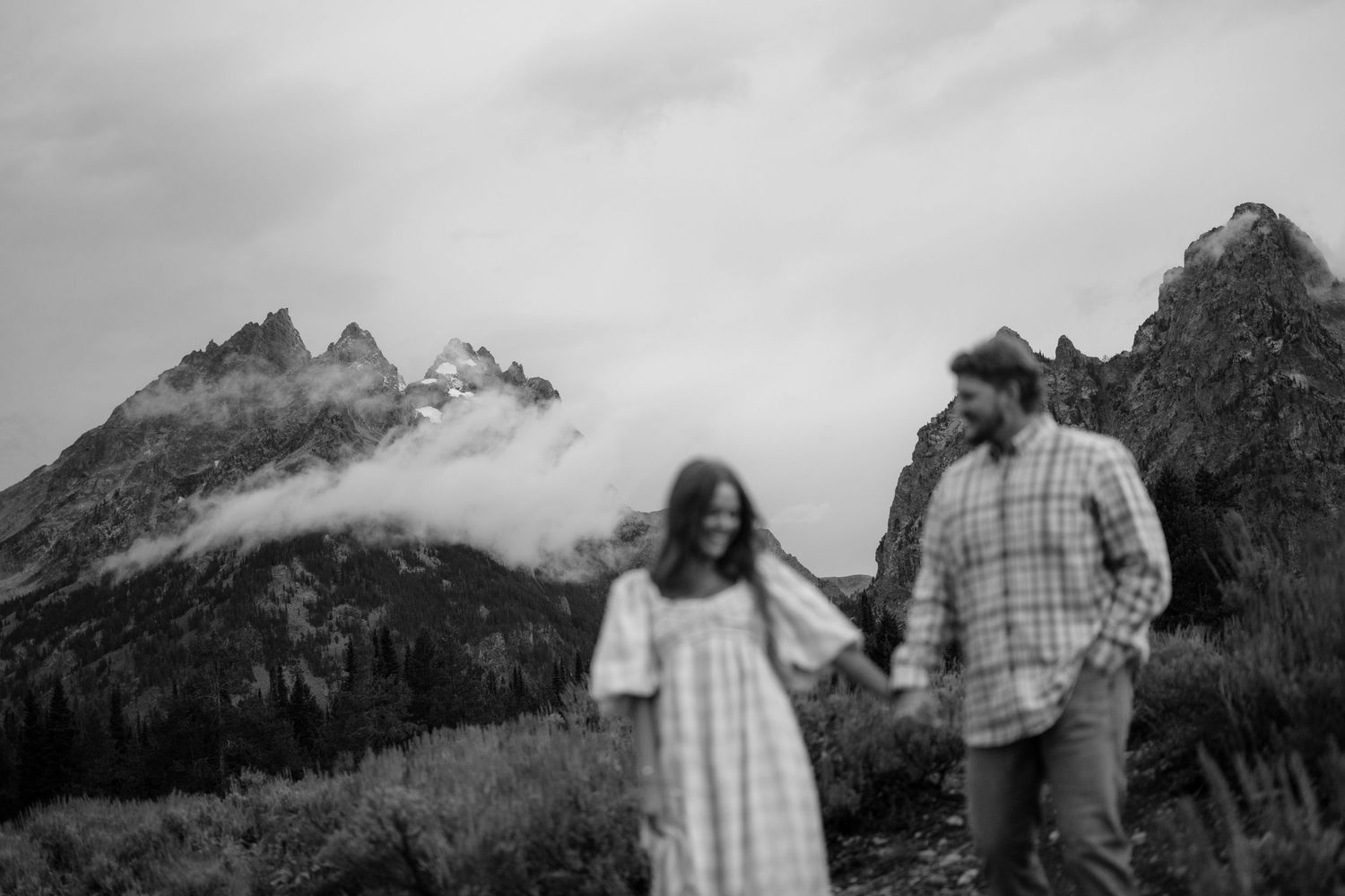 A couple holds hands as they walk through a mountainous, foggy landscape.