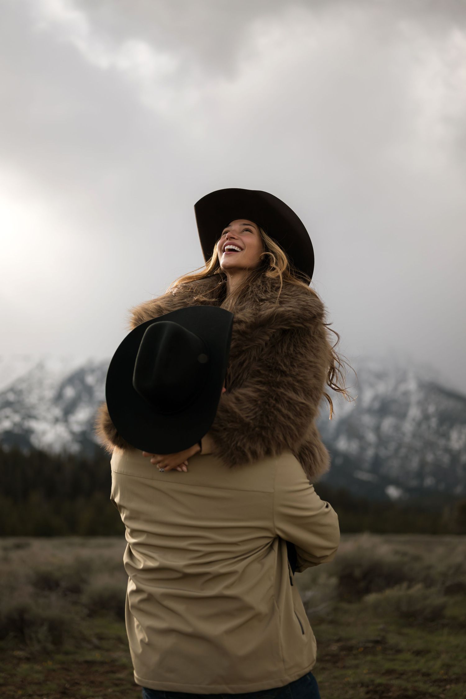 A joyful woman in a black cowboy hat and fur stole being lifted by a man outdoors with snowy mountains behind.