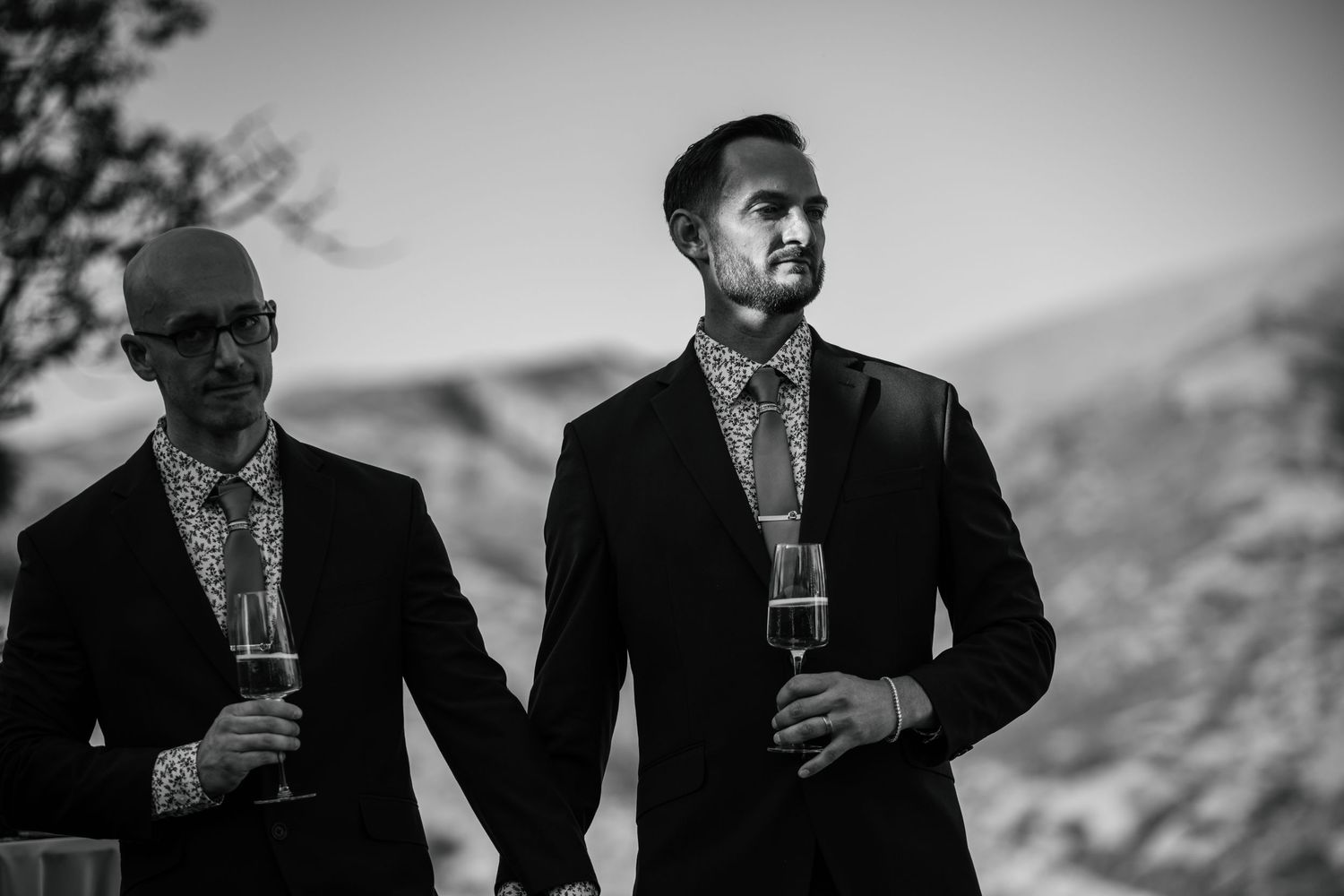 Two men in suits stand outdoors in a mountainous setting.