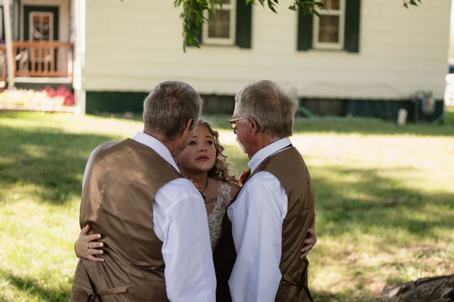 A bride embraced by two men in brown vests outdoors near a white house during a wedding ceremony.