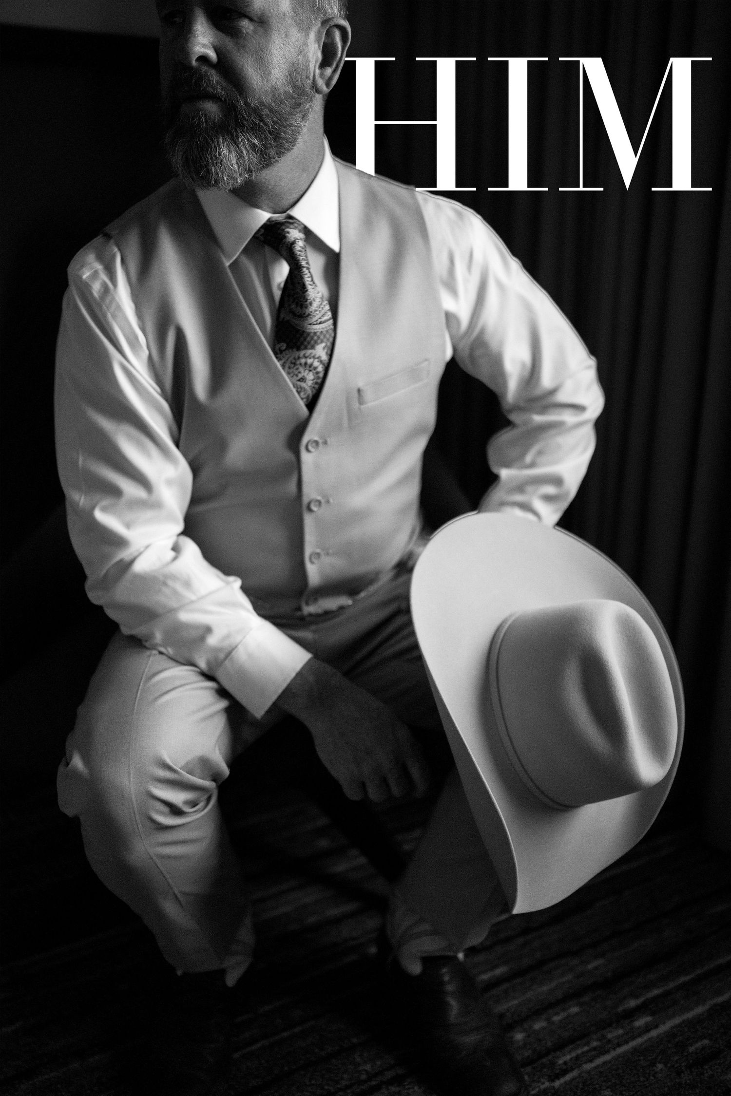 Black and white photo of a bearded man in a vest and tie holding a cowboy hat, styled for a masculine fashion editorial.