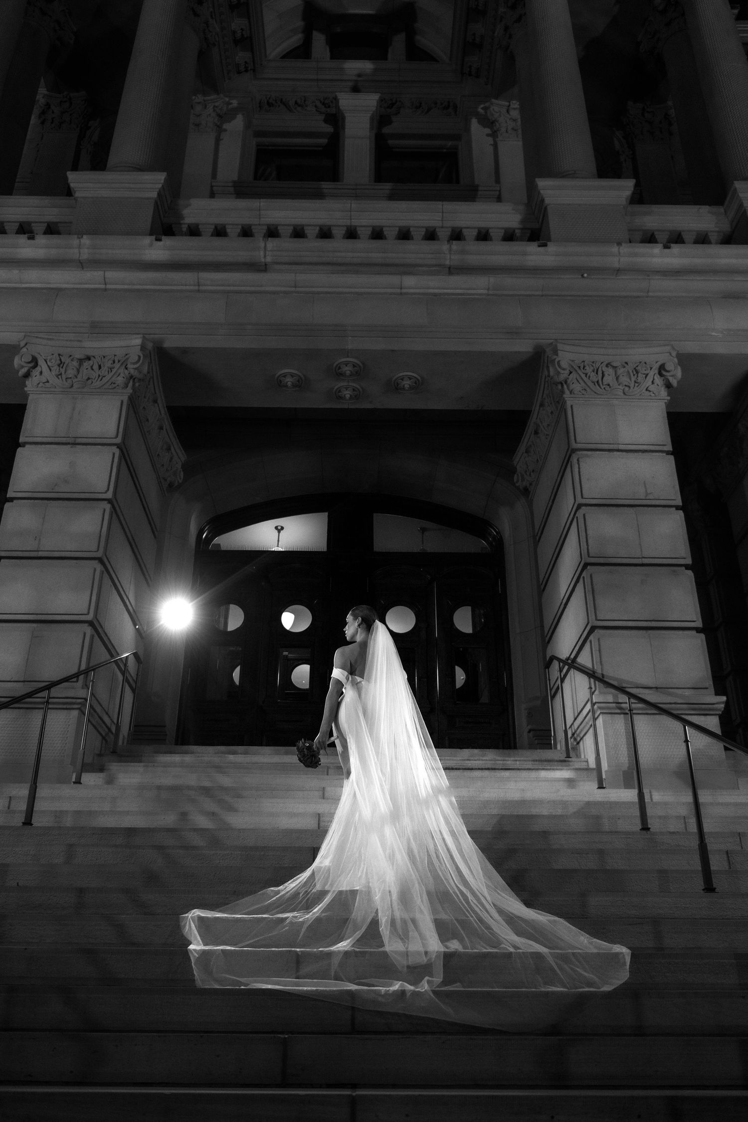 A bride in a long, flowing white wedding dress stands in front of a grand, ornate building at night.