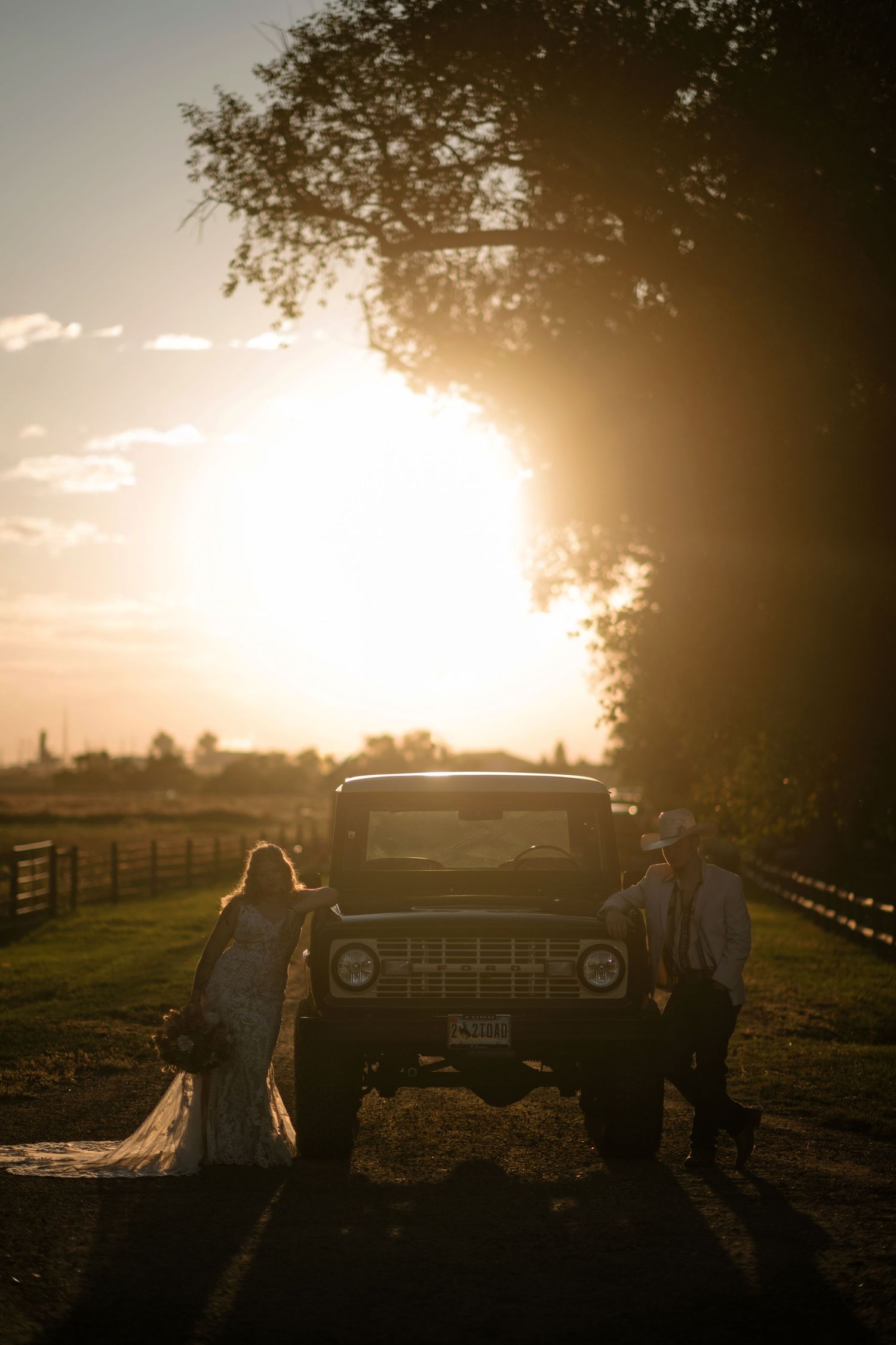 Bride and groom silhouetted beside a vintage truck at golden sunset on a rustic farm road.