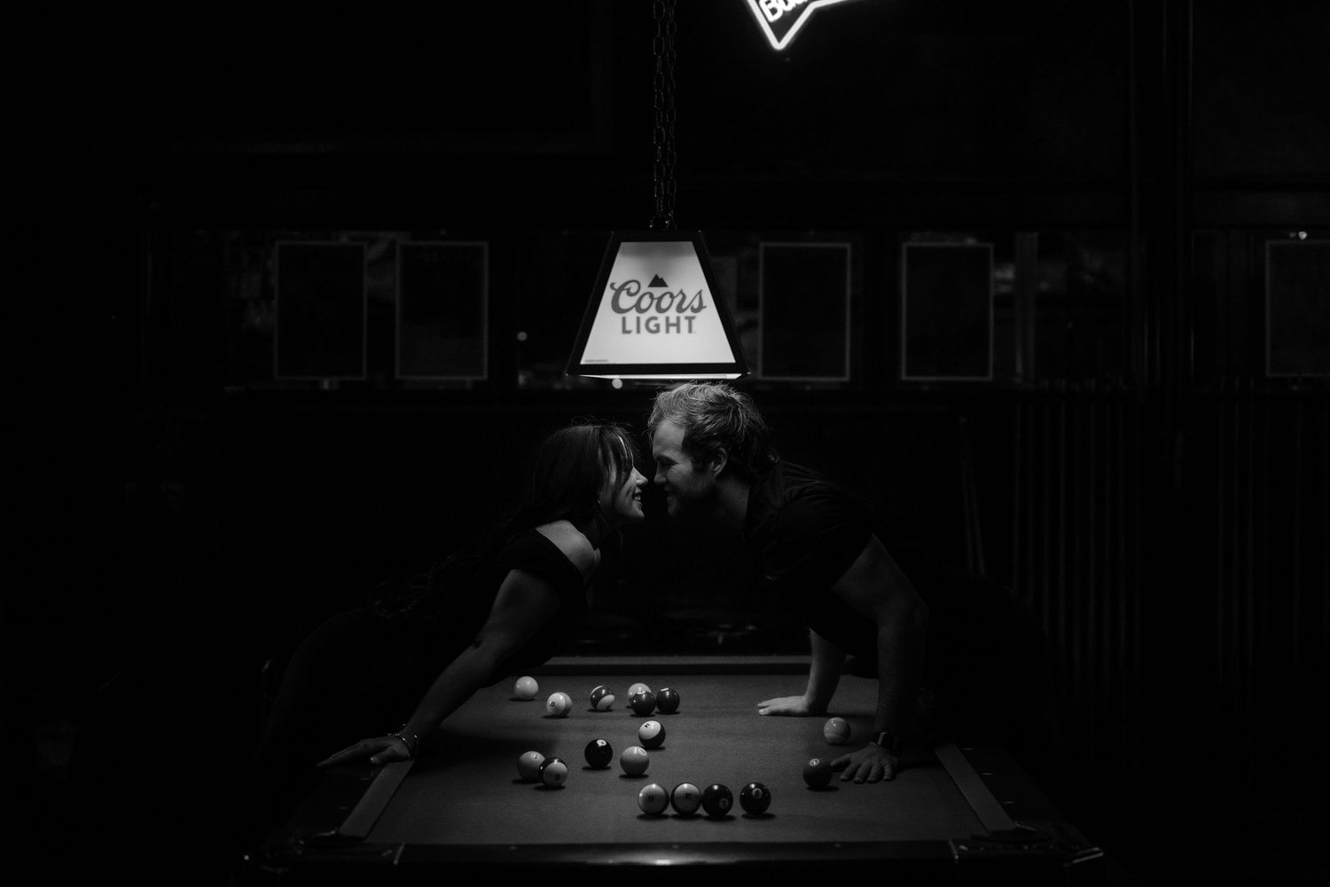 Couple sharing a romantic moment over a pool table under a Coors Light lamp in a dark bar, black and white photo.