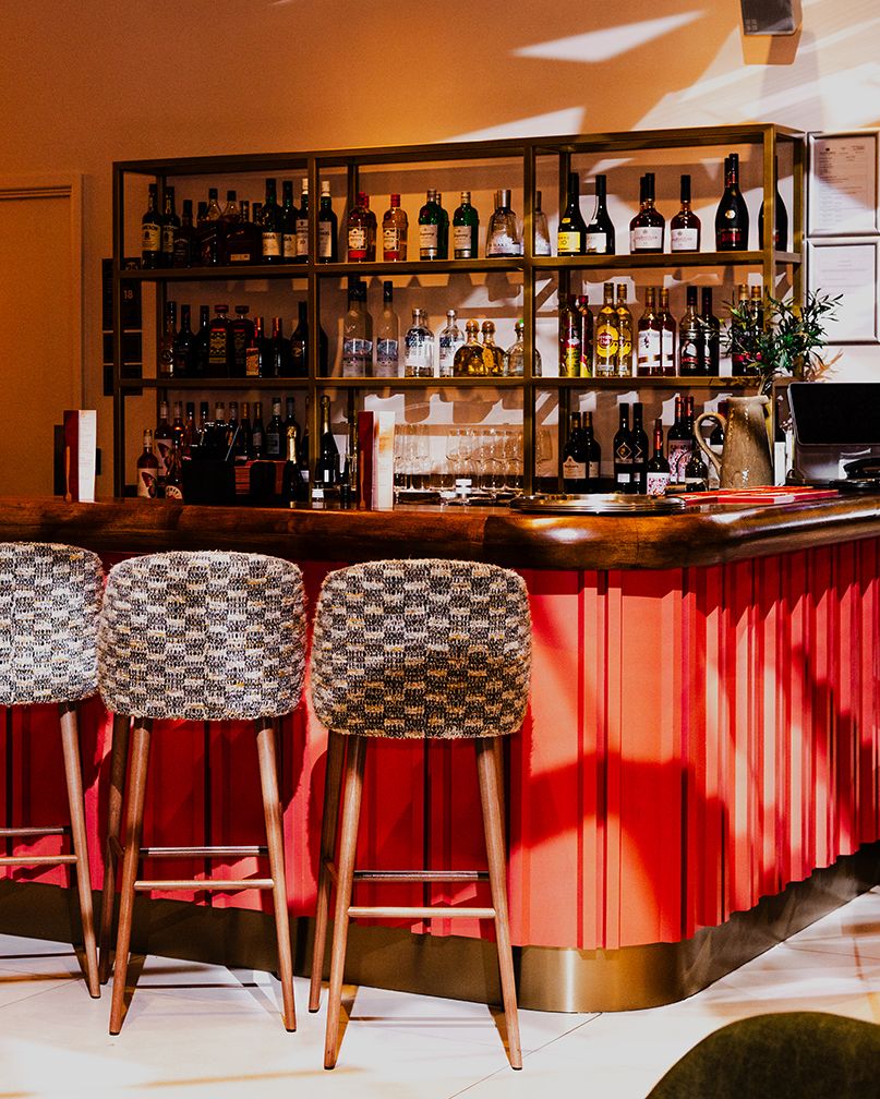 Modern bar interior with red corrugated metal base, wooden counter, and shelves of liquor bottles against warm lighting.