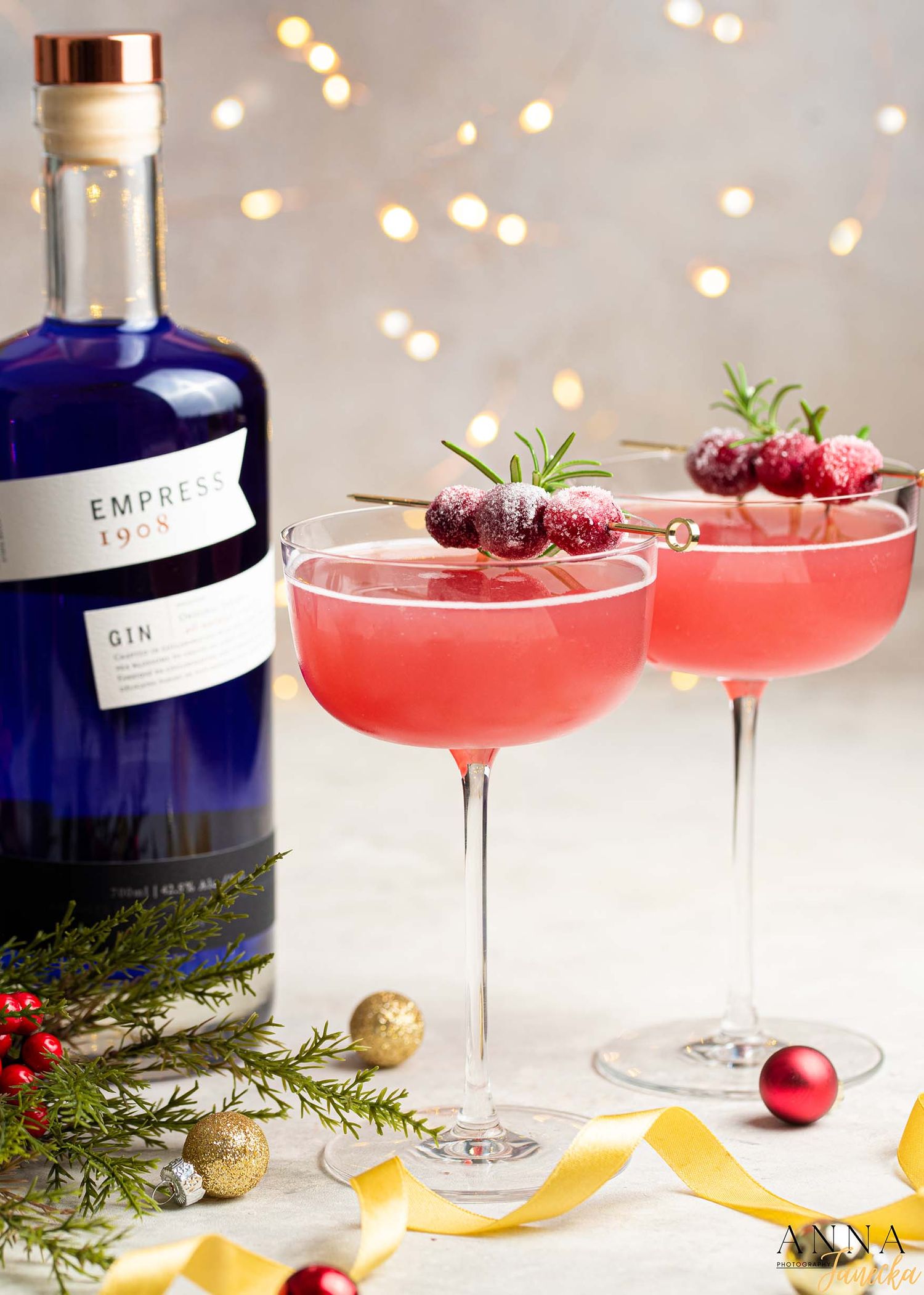 Pink gin cocktails garnished with fresh cherries and rosemary next to blue gin bottle.