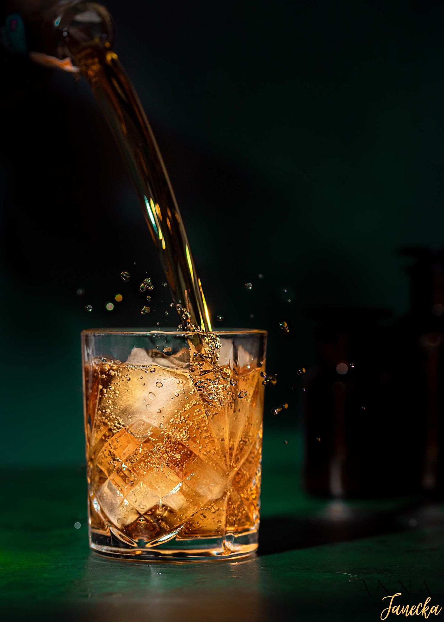 Amber-colored cocktail being poured over ice in a rocks glass against a dark green background.