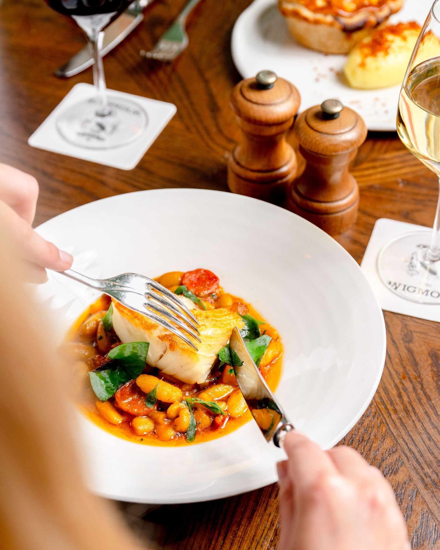 Person cutting into a pan-seared fish fillet with vegetables and beans in a white bowl at a restaurant table.