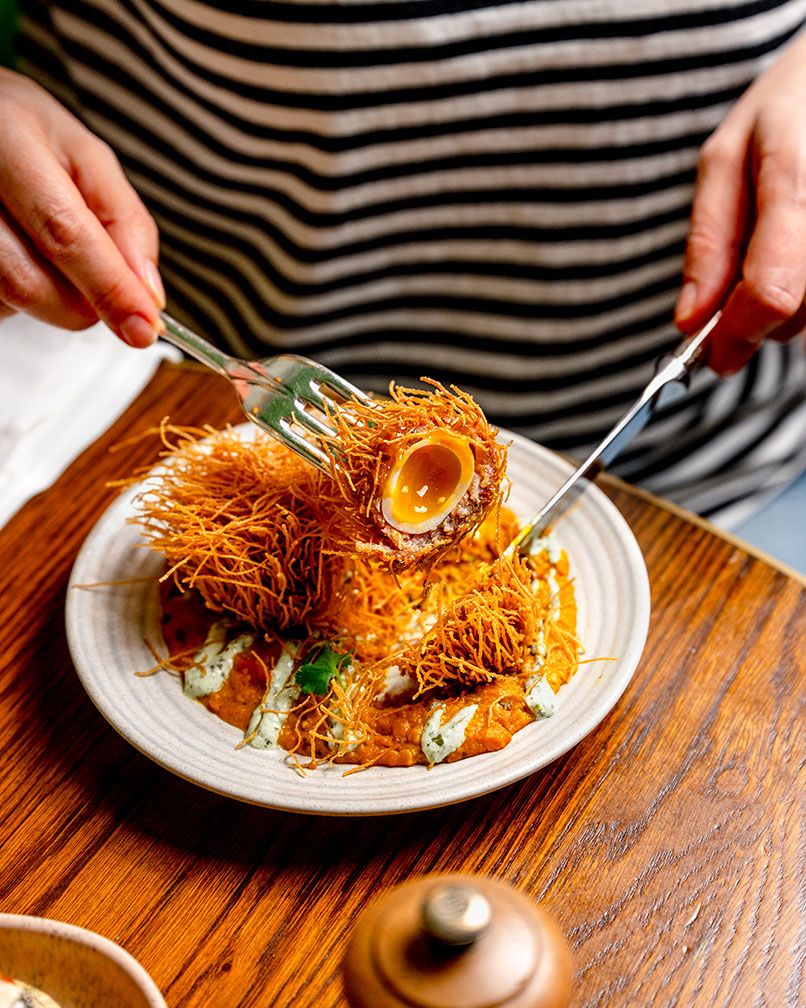 Person cutting into a crispy fried scotch egg with shredded potato crust on a sauced plate at a restaurant.