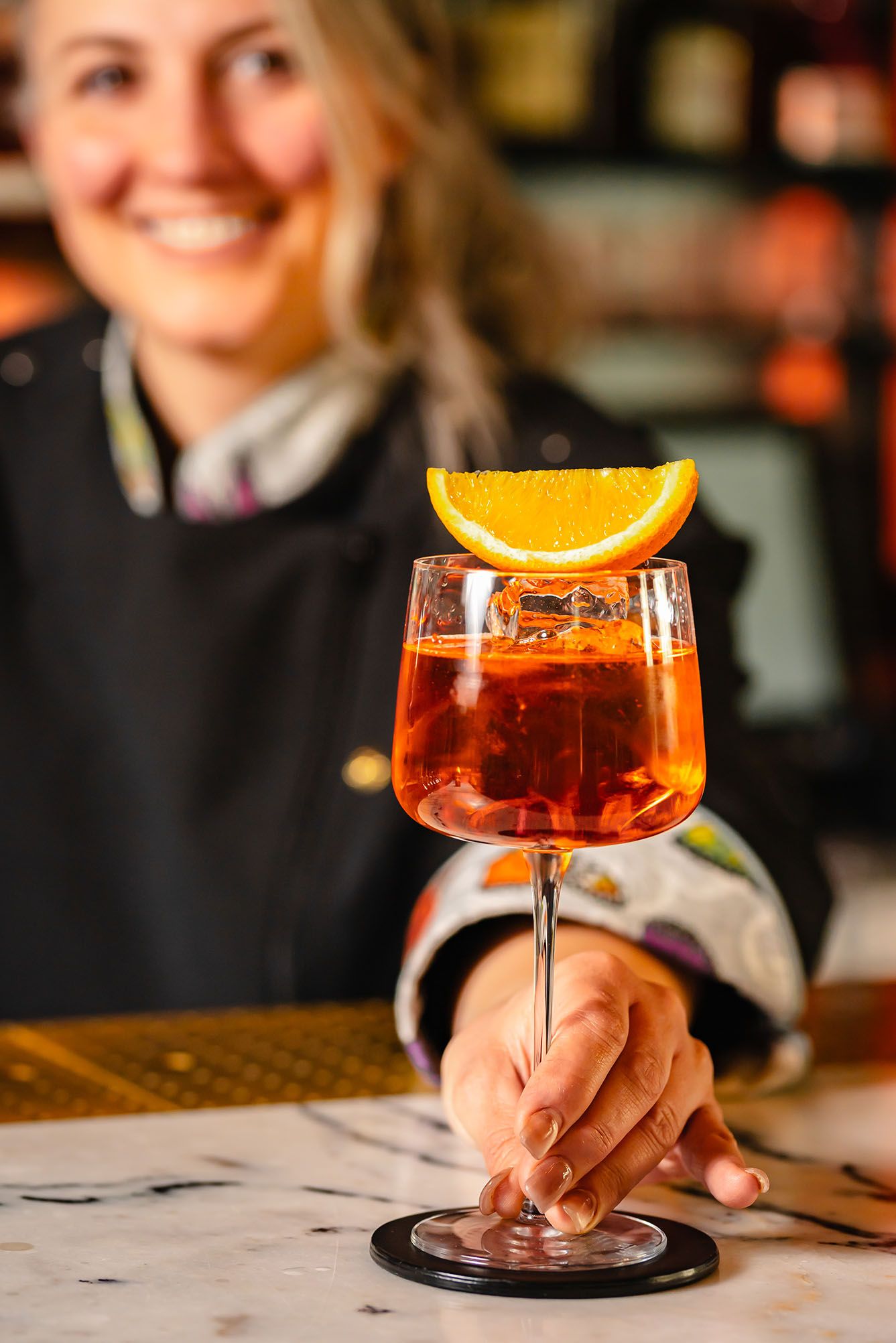 Smiling bartender presenting a vibrant Aperol Spritz cocktail garnished with an orange slice on a marble bar.