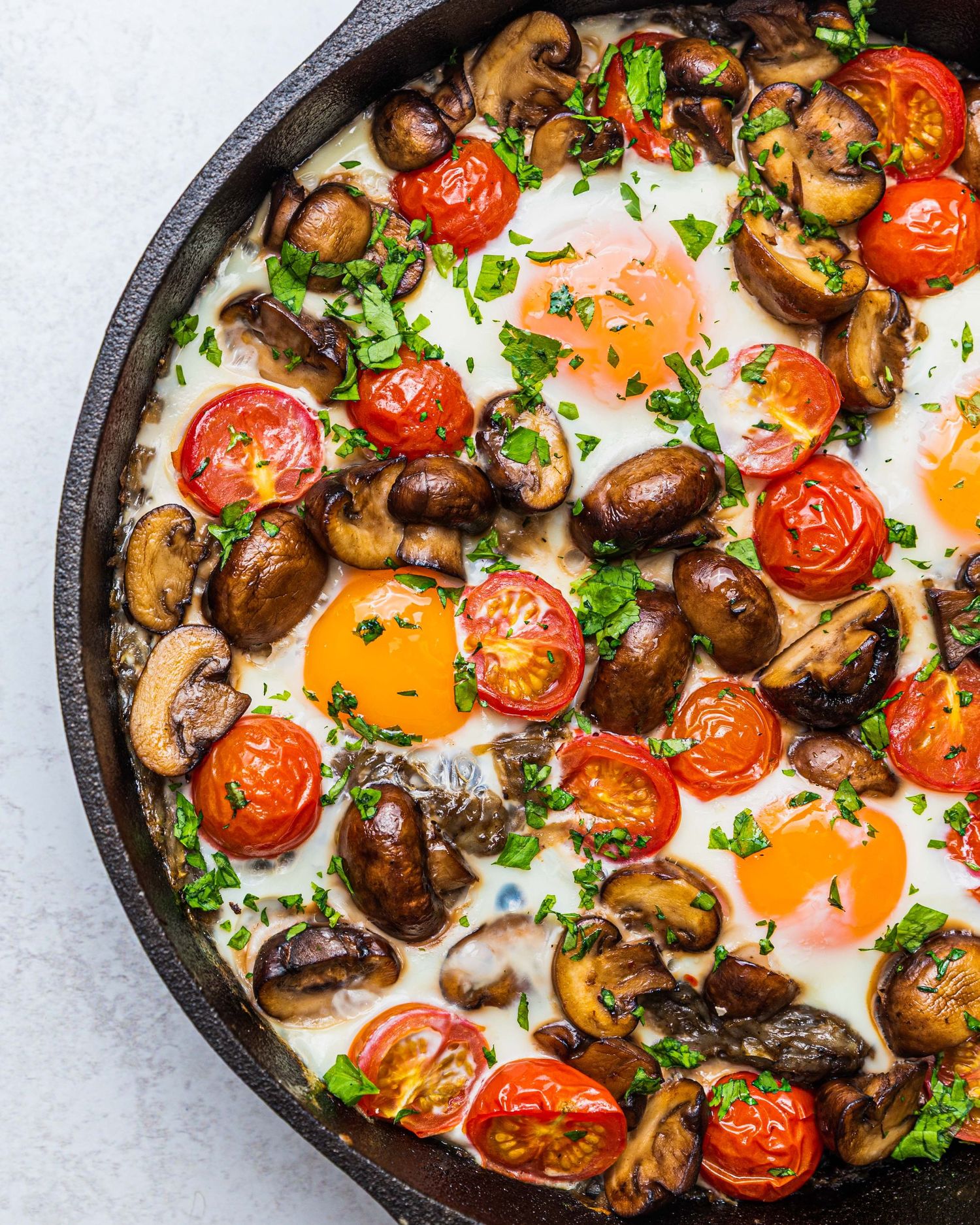 Cast iron skillet with baked eggs, sautéed mushrooms, roasted cherry tomatoes, and fresh parsley for a hearty breakfast.