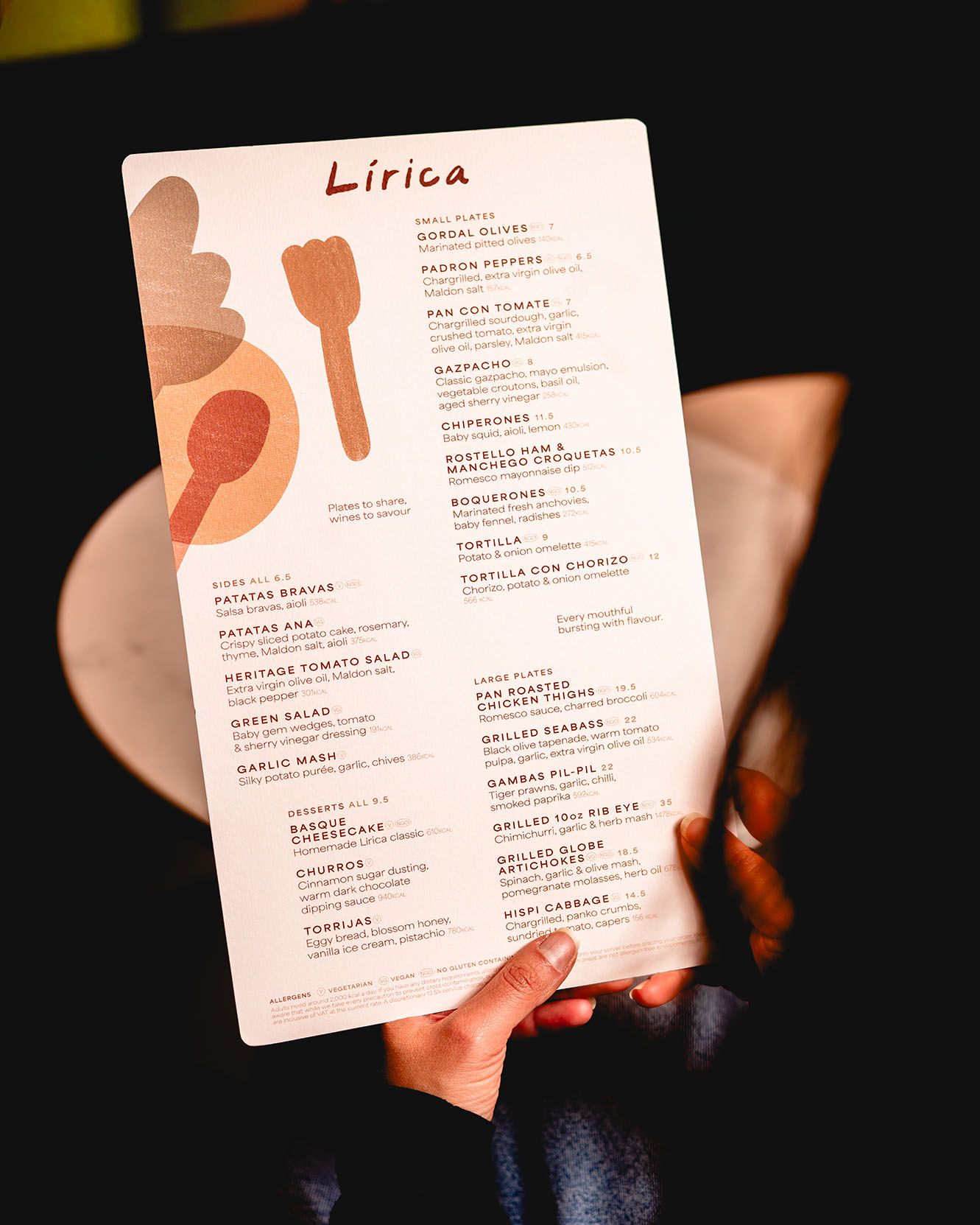 Hand holding Lirica restaurant menu featuring Spanish small plates, tapas, and sides with decorative utensil graphics.