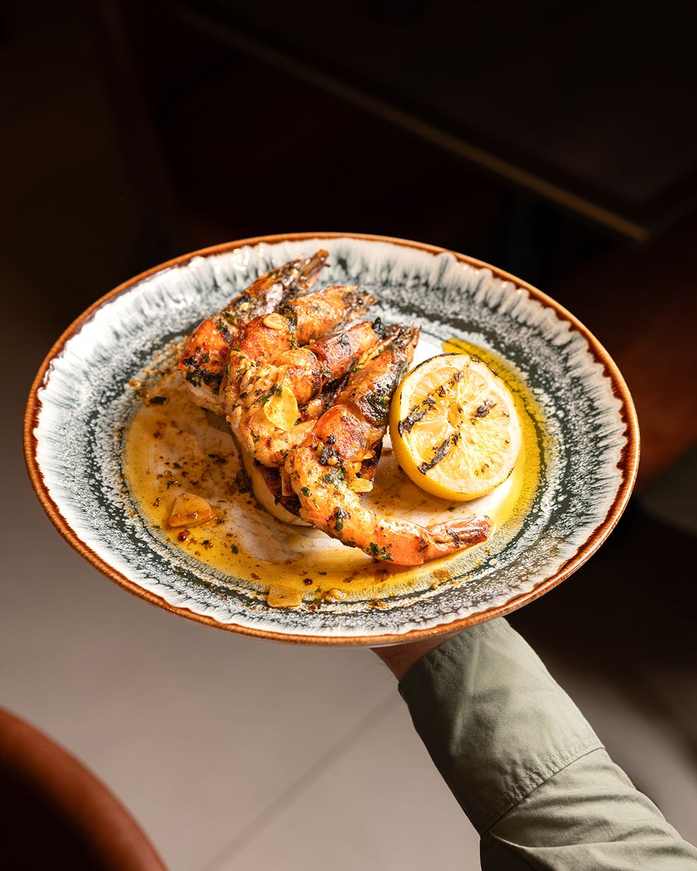 A gloved hand holds up a decorative plate with grilled shrimp and lemon slices in a dark moody setting.