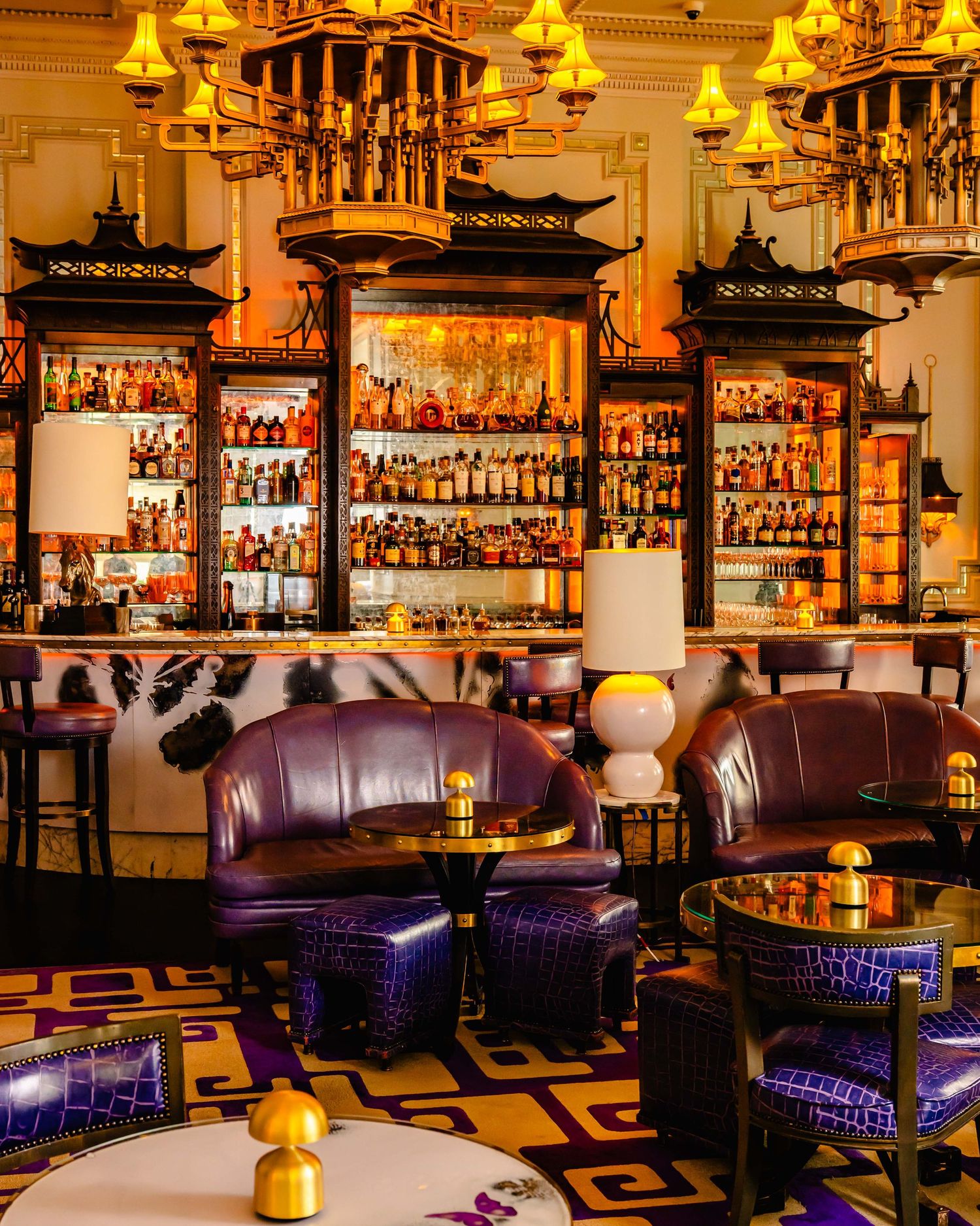 Opulent bar lounge with purple velvet seating, gold chandeliers, and backlit shelves of spirits in an Asian-inspired decor.
