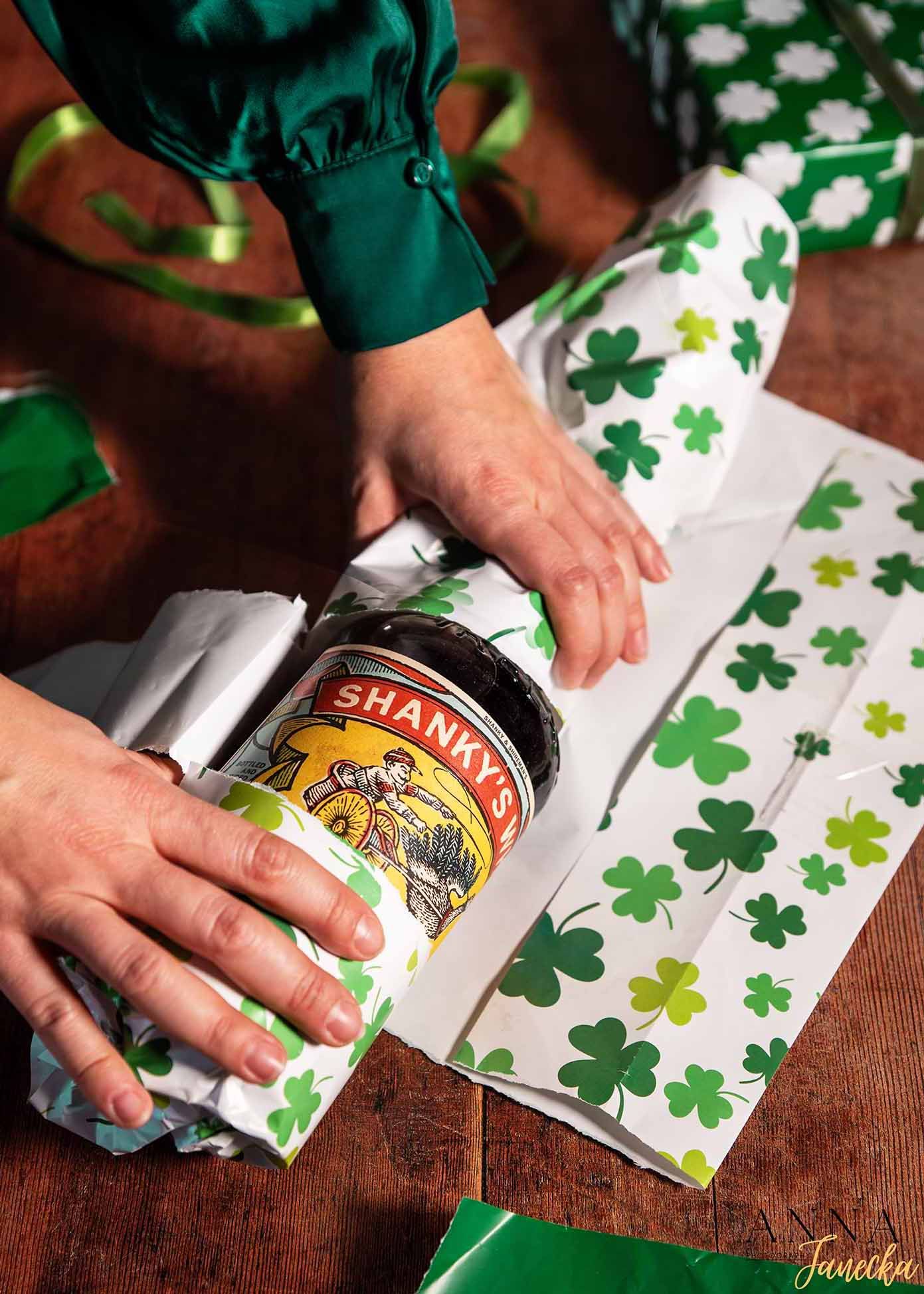 Hands holding a bottle of Shandy beer against a festive shamrock-patterned background for St. Patrick's Day.