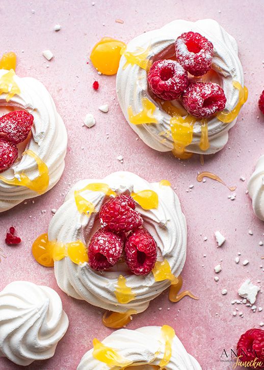 Mini pavlovas topped with fresh raspberries and orange curd on a pink surface.