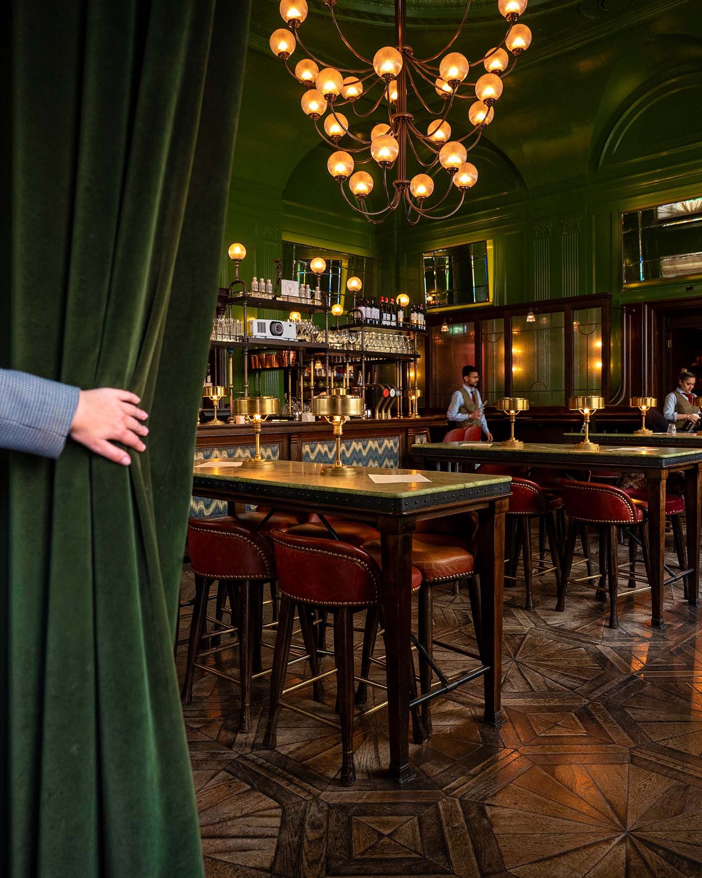 Elegant green velvet bar interior with warm globe chandelier, red leather stools, and ornate parquet flooring.