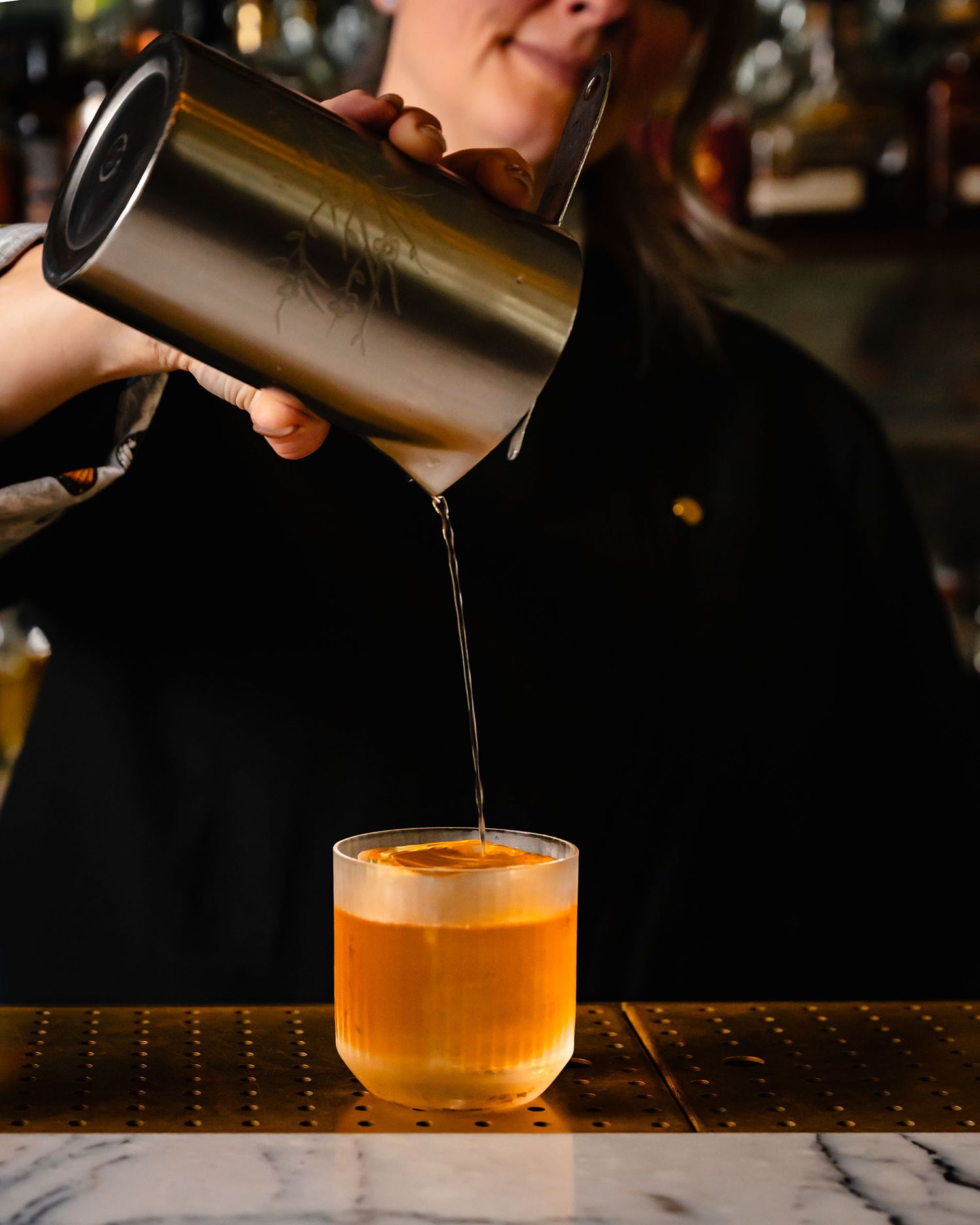 Bartender pouring a golden cocktail from a shaker into a rocks glass on a marble bar counter.